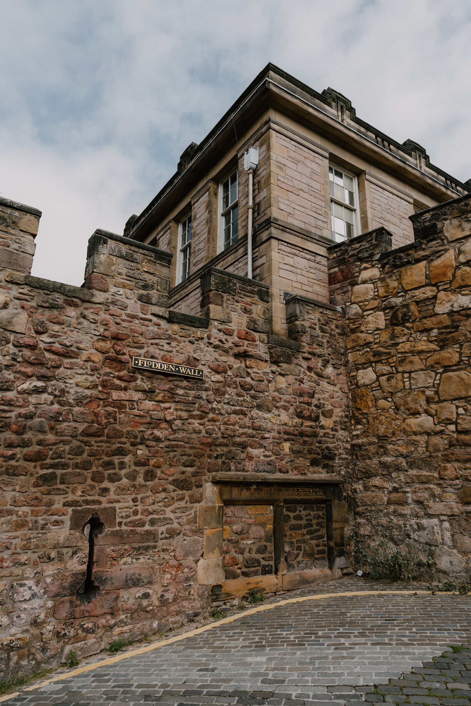 A sign reading 'Flodden Wall' is fixed to a stone rubble wall beneath a sandstone tenement corner.
