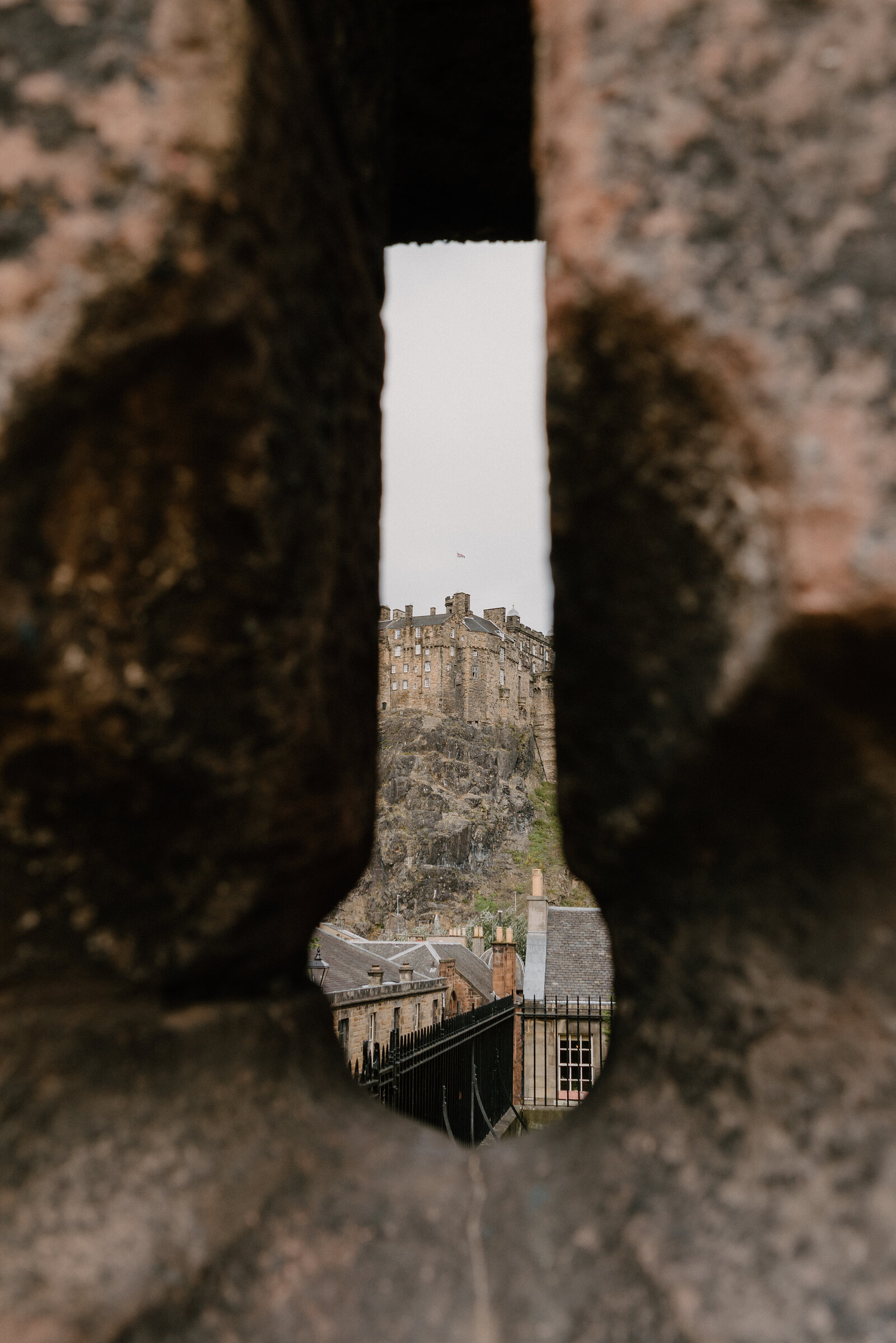 Edinburgh Castle and rooftop chimneys are framed through a narrow gap in a stone parapet wall.