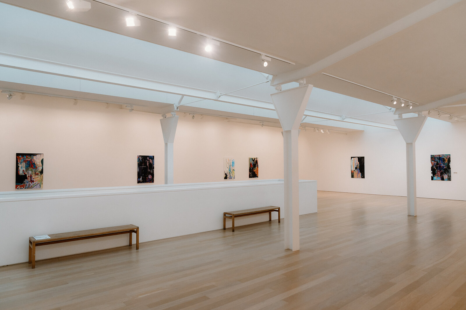 A row of framed artworks lines a white gallery wall beneath skylights, wooden benches set along a raised walkway.
