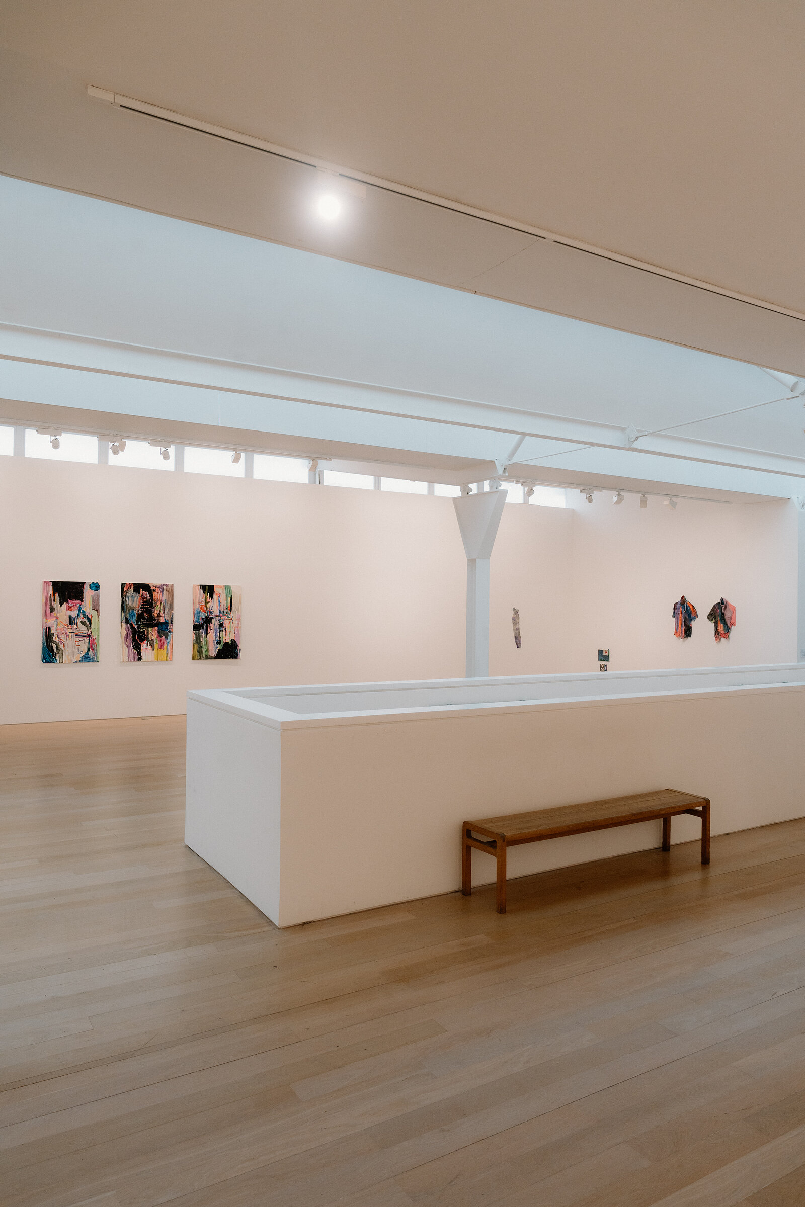 A white plinth stands in the centre of a gallery room hung with small paintings, a wooden bench nearby.