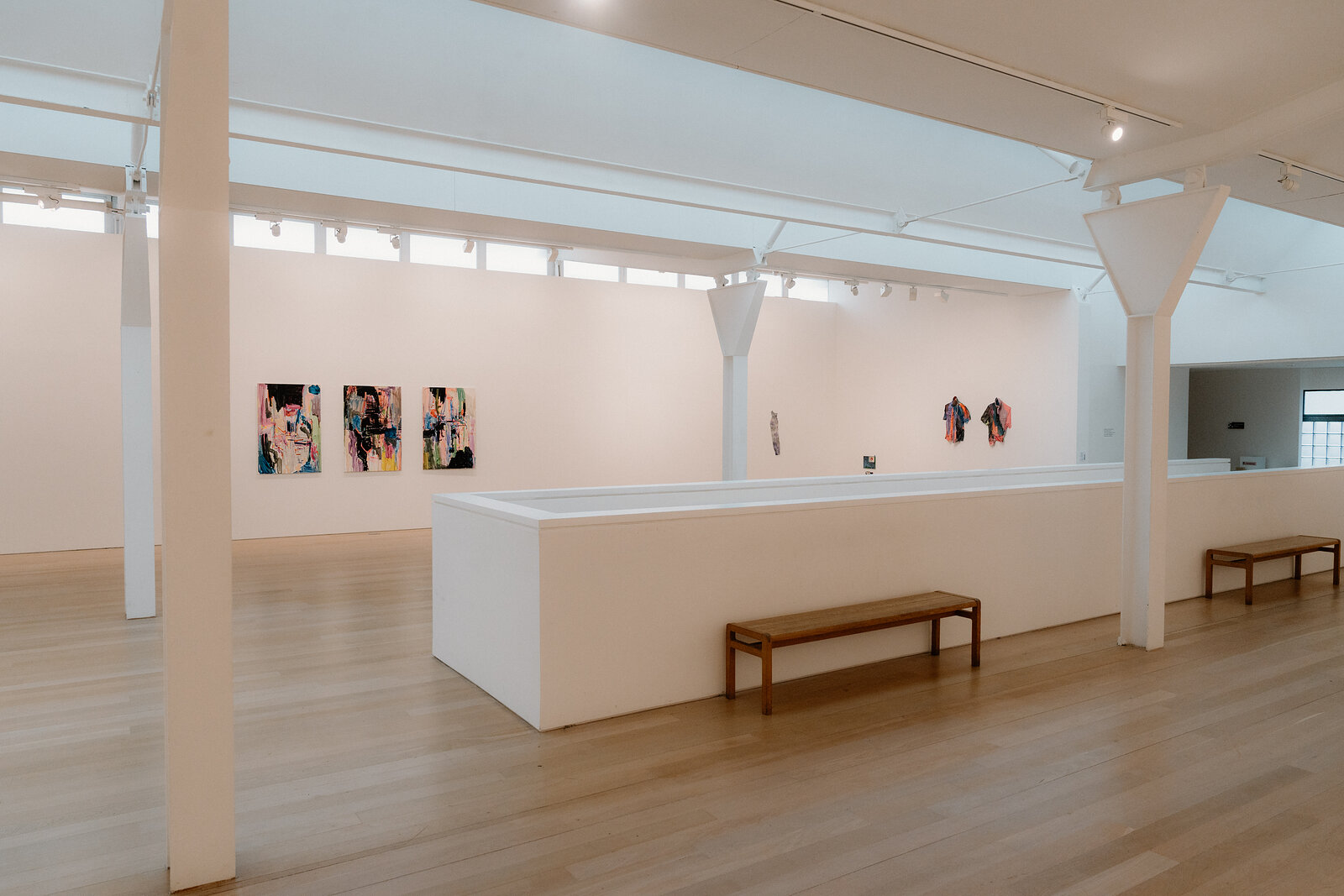 Framed paintings line a gallery wall beside a long white plinth and wooden bench beneath skylights.