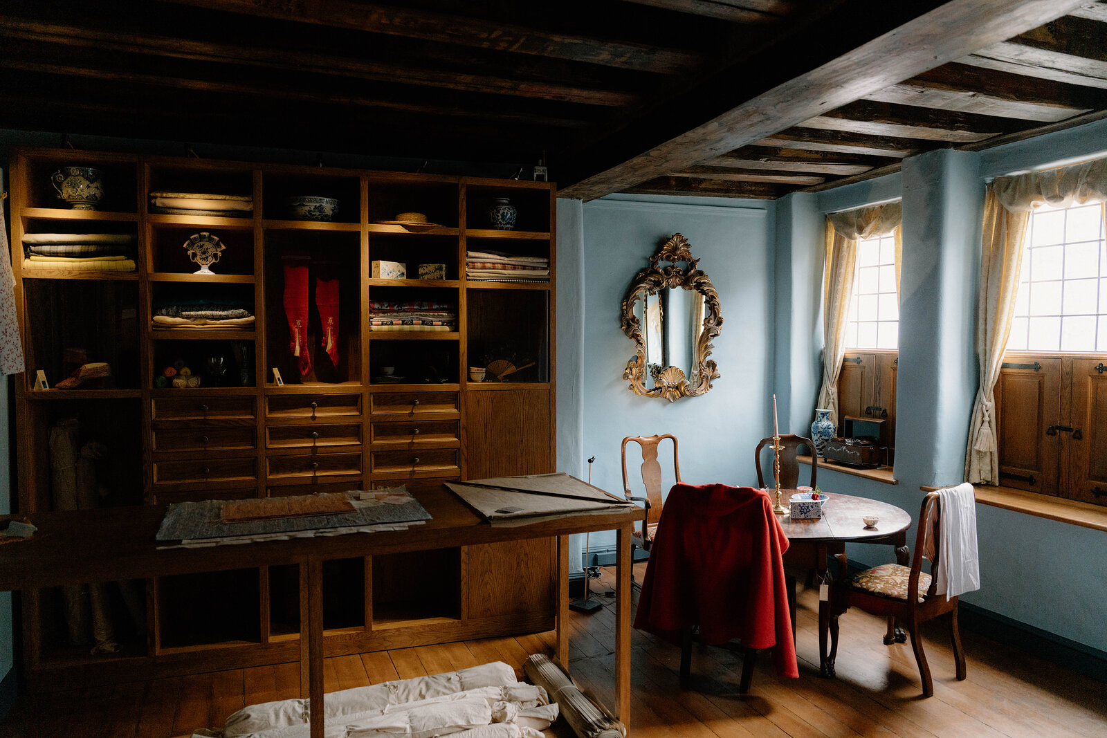 A wooden bureau desk with china ornaments stands beside a red-draped chair in a blue-walled period room with beamed ceilings.