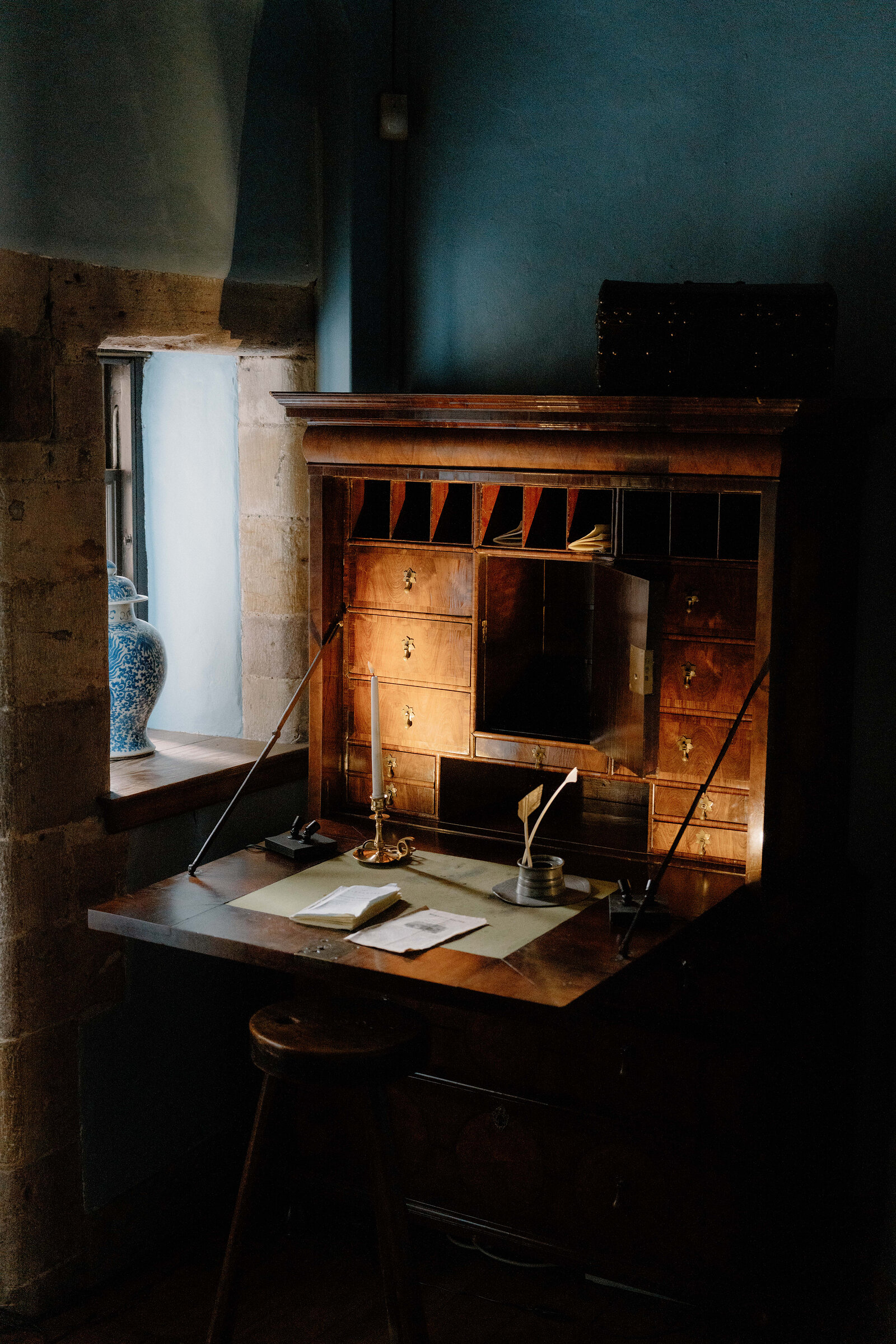 An antique writing bureau with pigeonholes and drawers stands open beside a stone-mullioned window, a candlestick and quill on the desk.