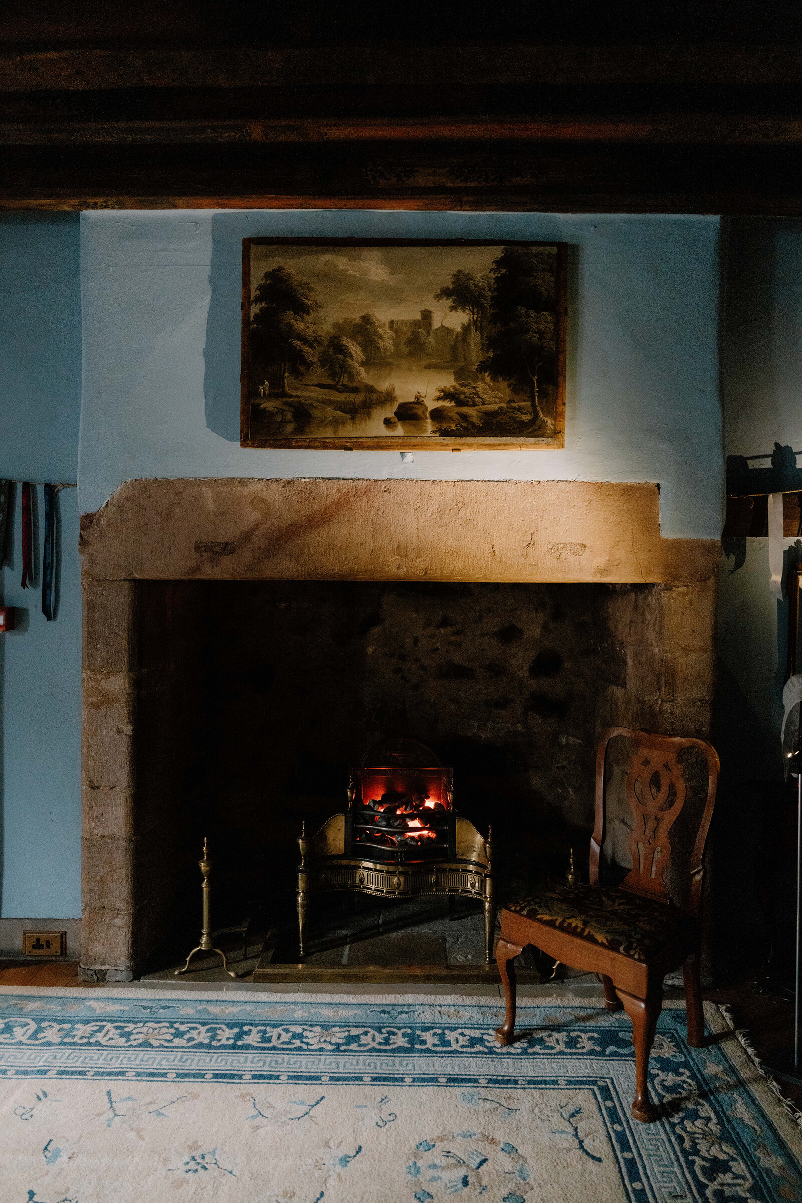 A landscape painting hangs above a small coal-burning fireplace flanked by an upholstered chair in a blue-walled period room.