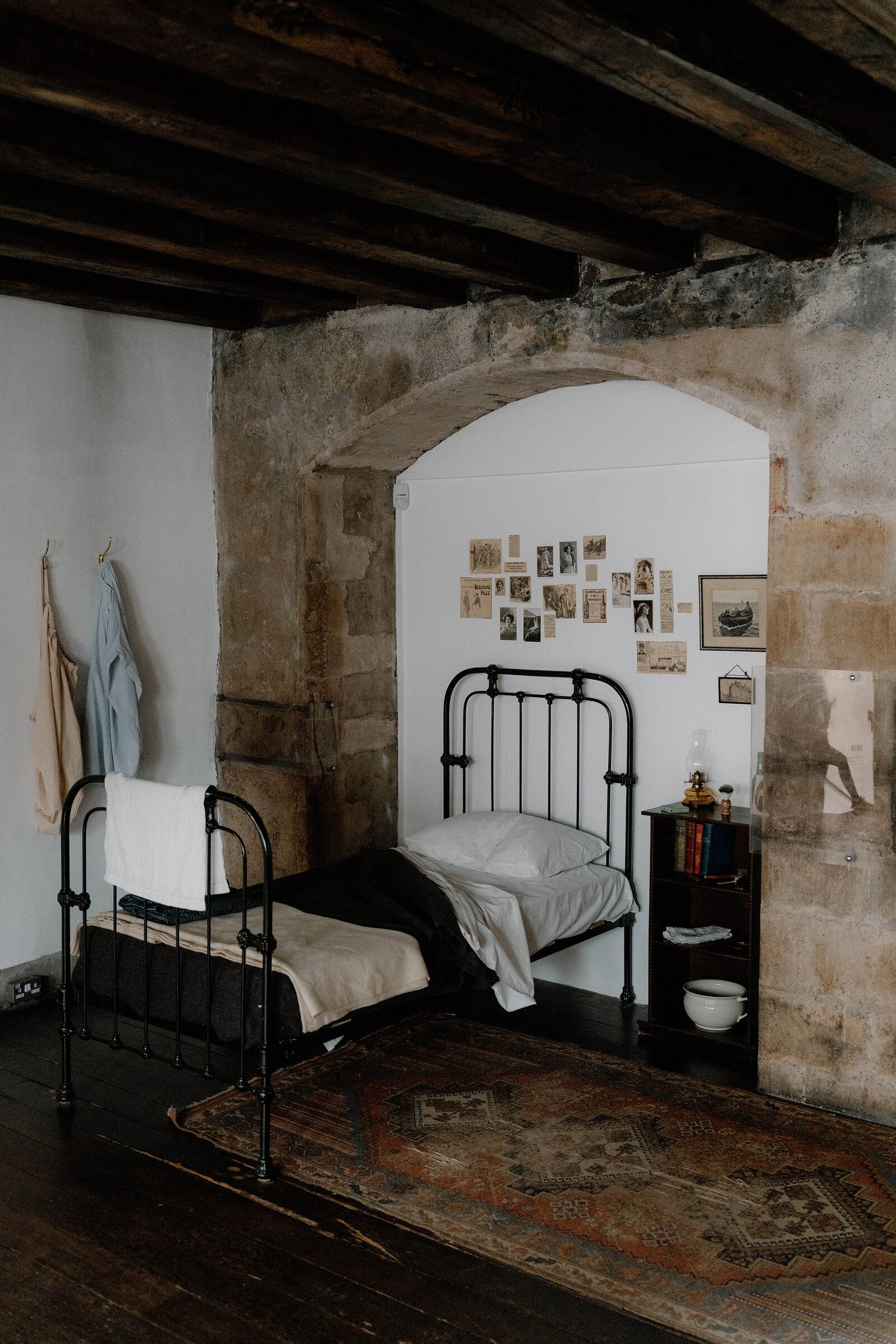An iron bed sits beneath a stone archway hung with small vintage photographs at Gladstone's Land, a wardrobe of coats nearby.