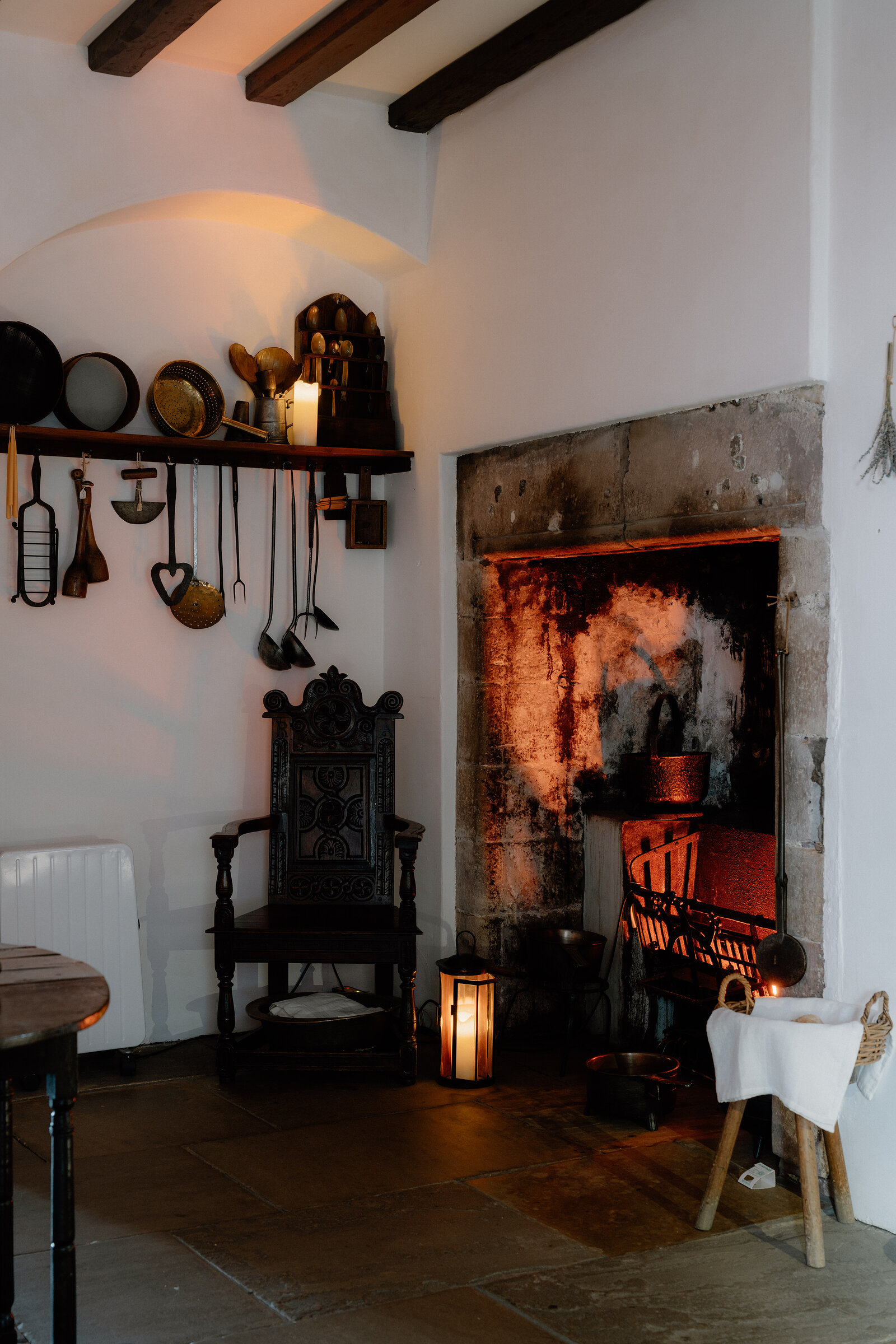 Copper pots and cooking utensils hang above a carved wooden chair beside a glowing stone fireplace at Gladstone's Land.