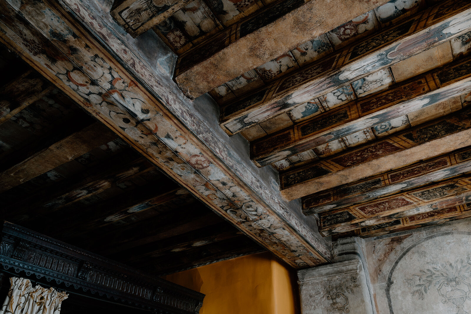 Faded decorative patterns cover a painted timber beam ceiling above a stone fireplace.