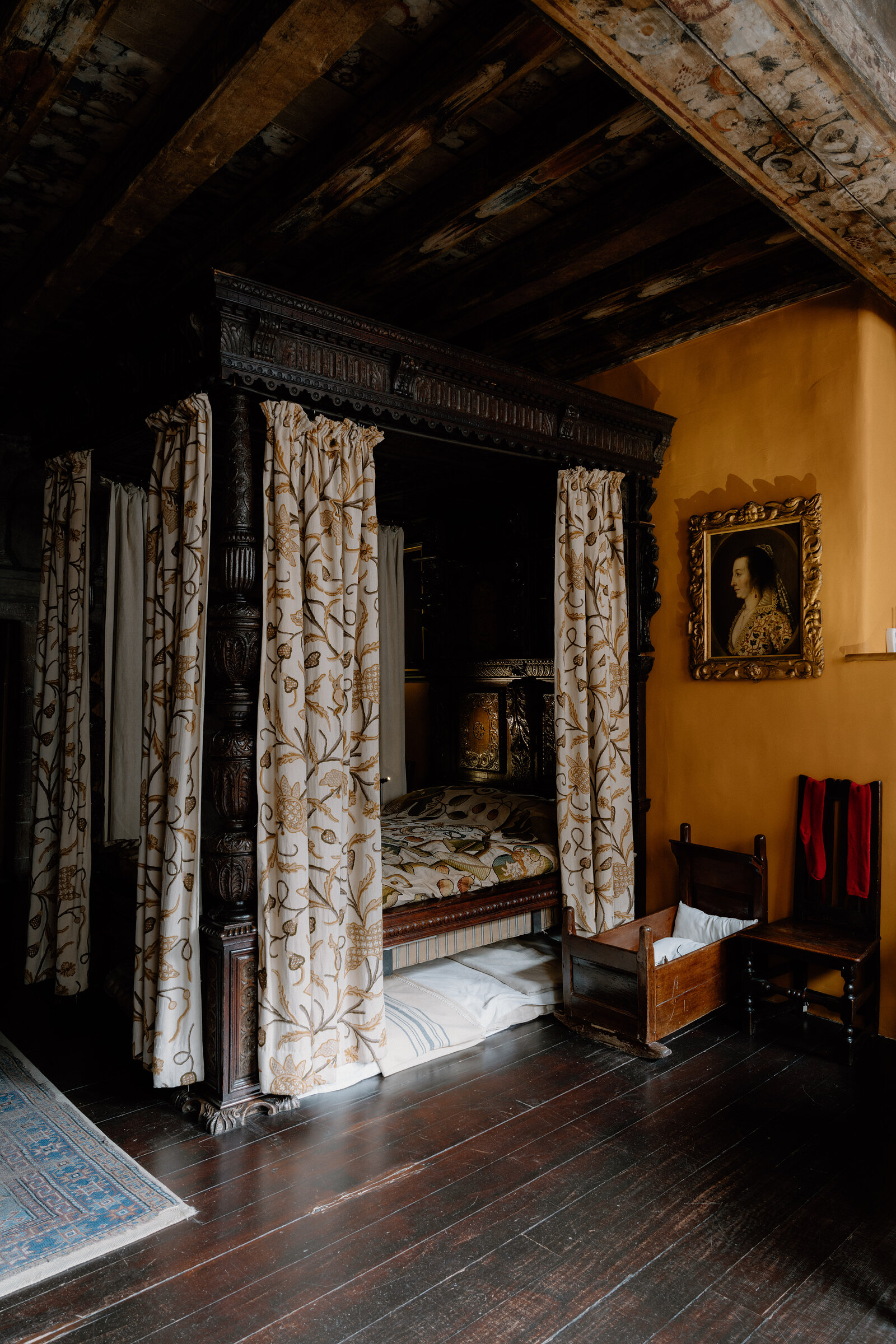 A carved oak four-poster bed with floral curtains stands opposite a wooden cradle and a framed portrait, beneath a painted beam ceiling.