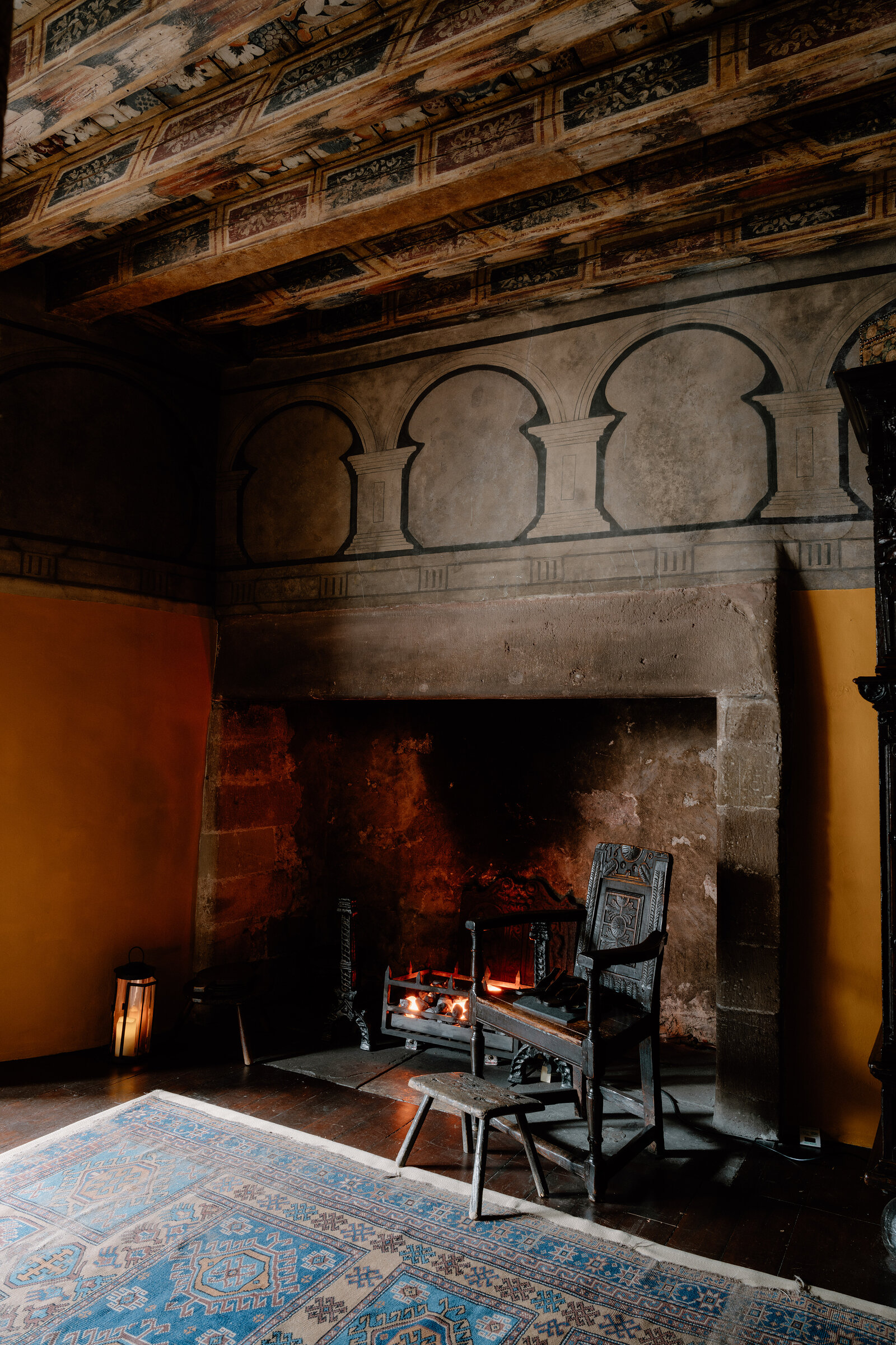 A carved wooden chair sits beside a lit fire in a stone hearth, beneath an arched painted frieze and a patterned rug.
