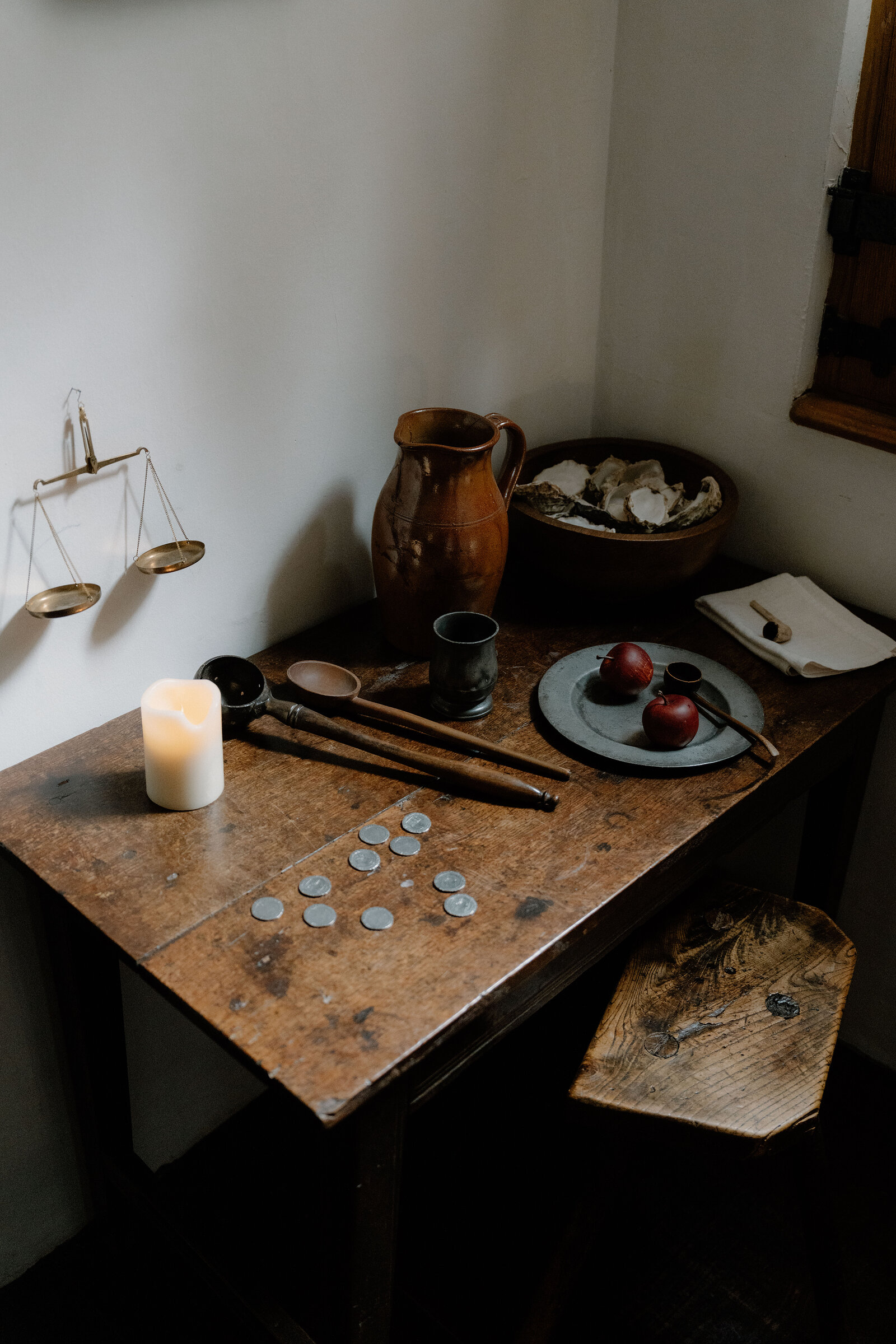 Brass scales, an earthenware jug, old coins and a plate of apples sit on a wooden table beside a lit candle in a historic room.