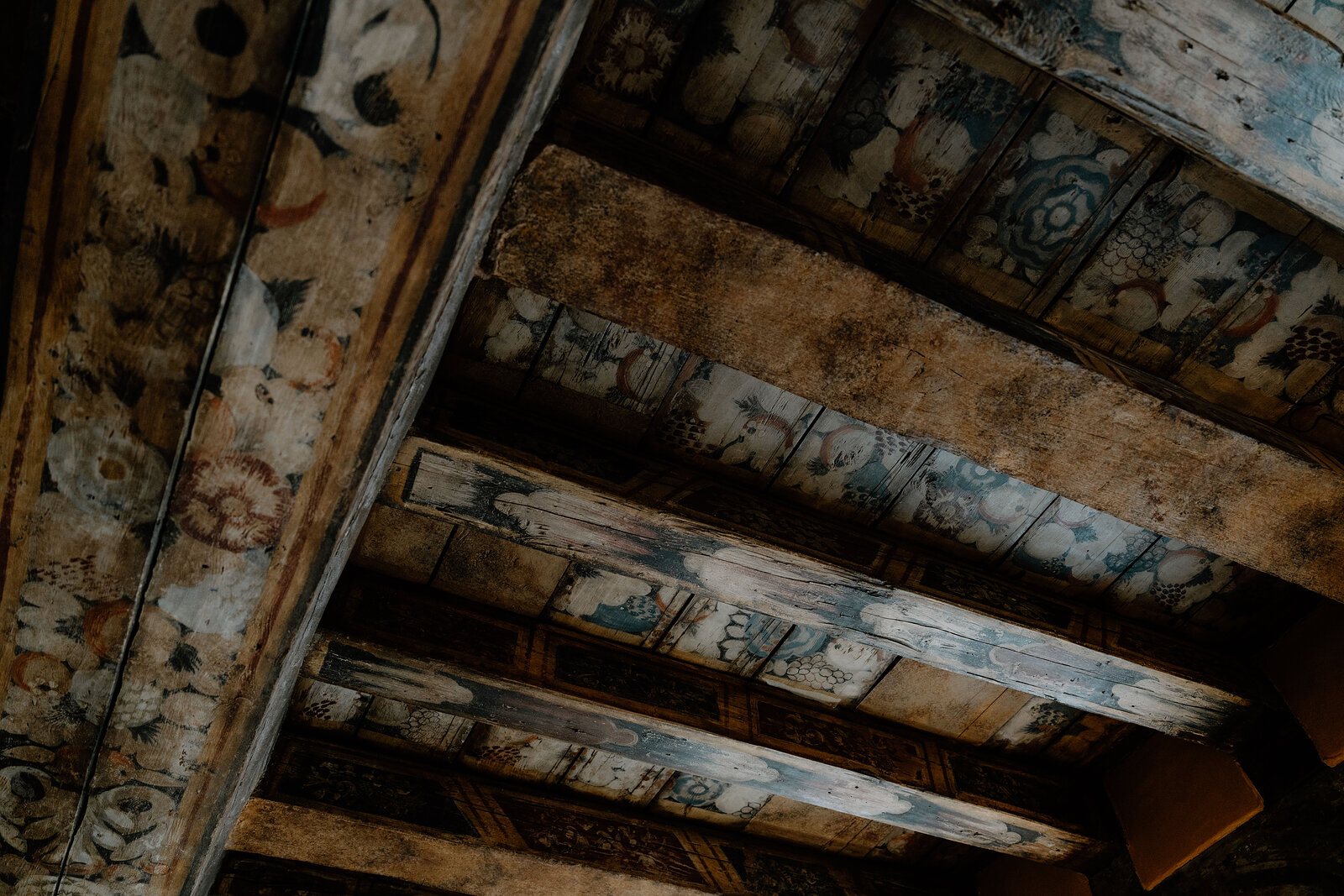 Faded decorative patterns cover a painted timber beam ceiling in a historic room.