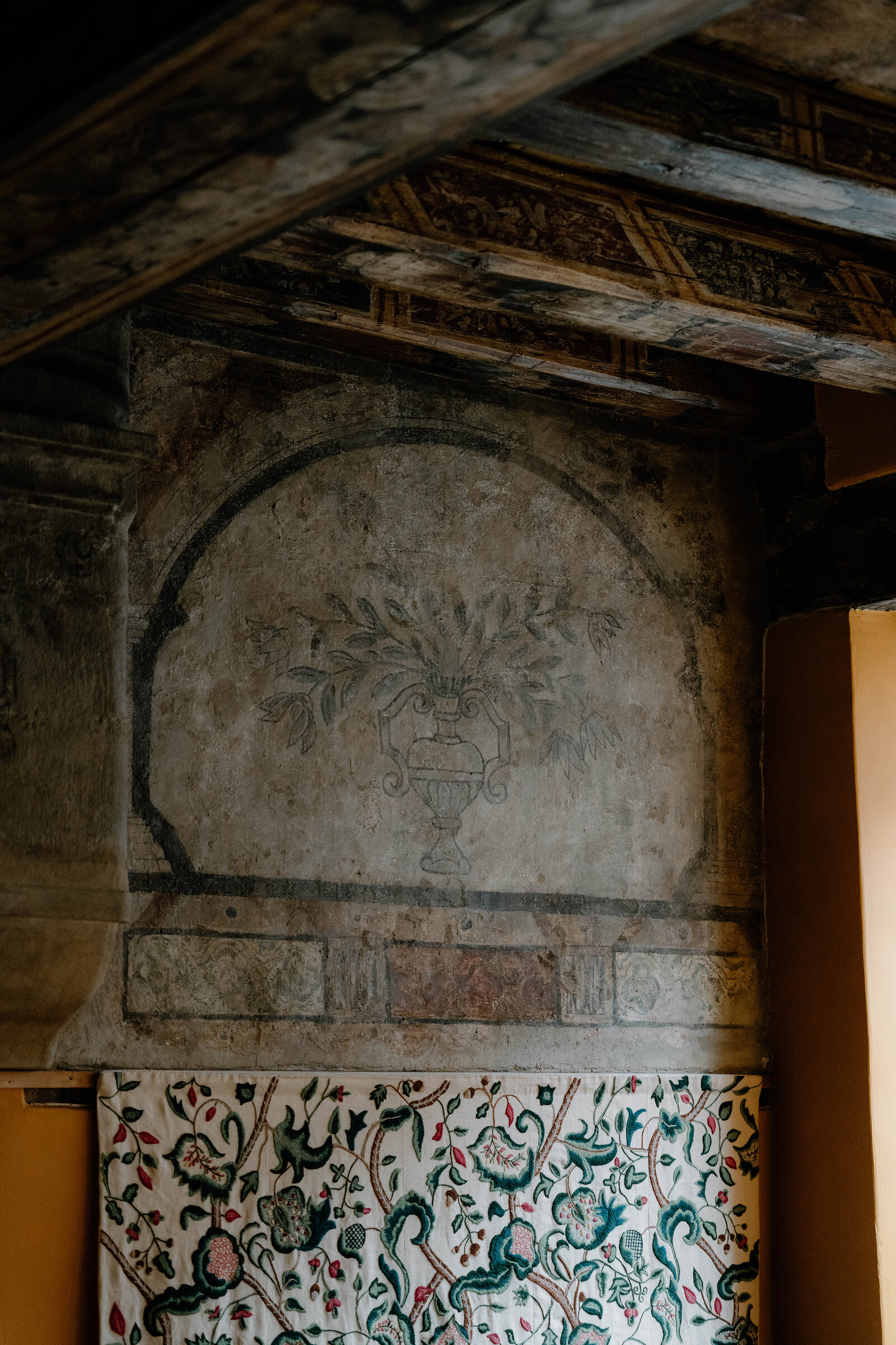 An arched painted frieze of an urn and flowers decorates the wall above a patterned tapestry in a historic room.