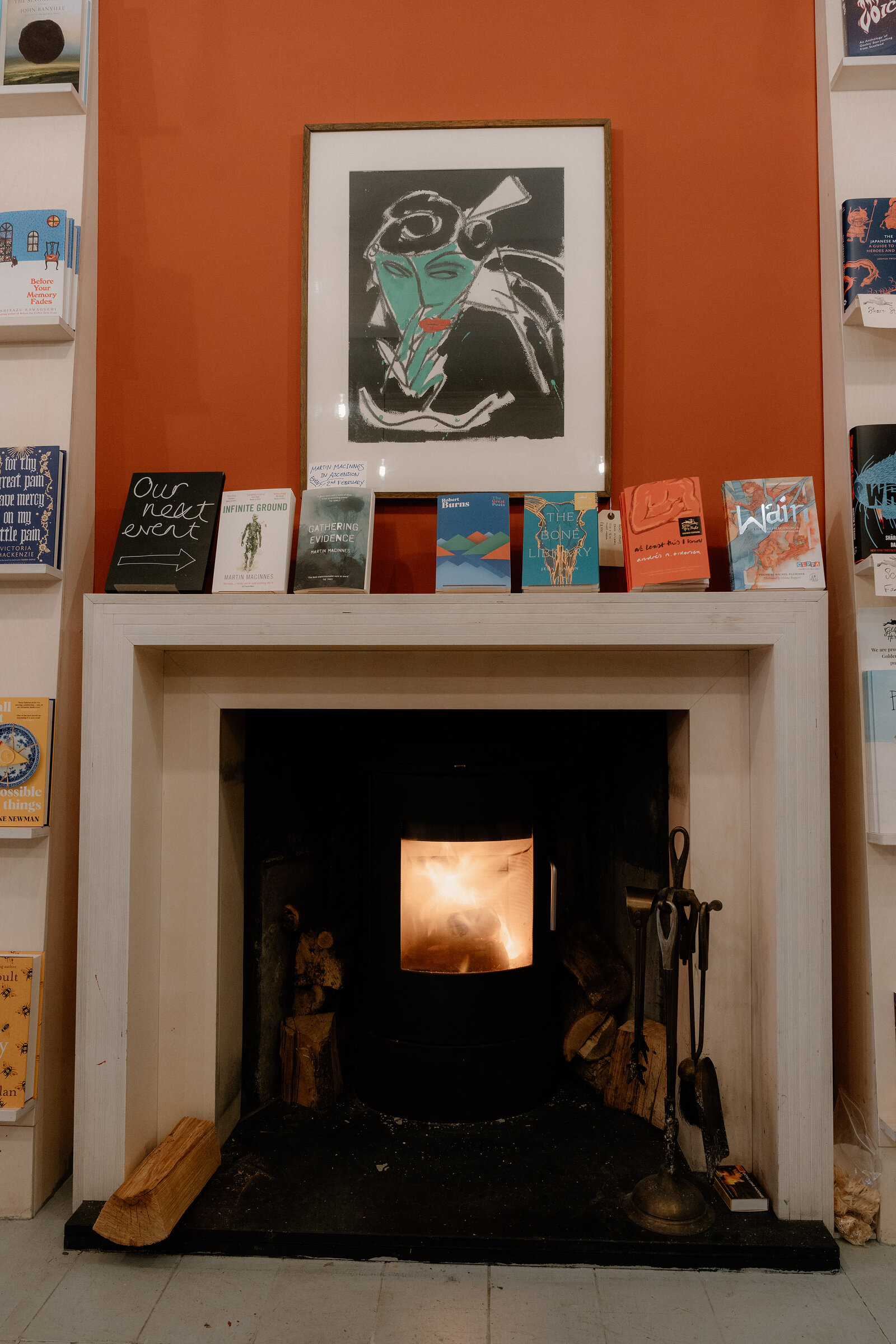 A framed illustration hangs above a mantel stacked with books and a lit stove at Golden Hare Books.