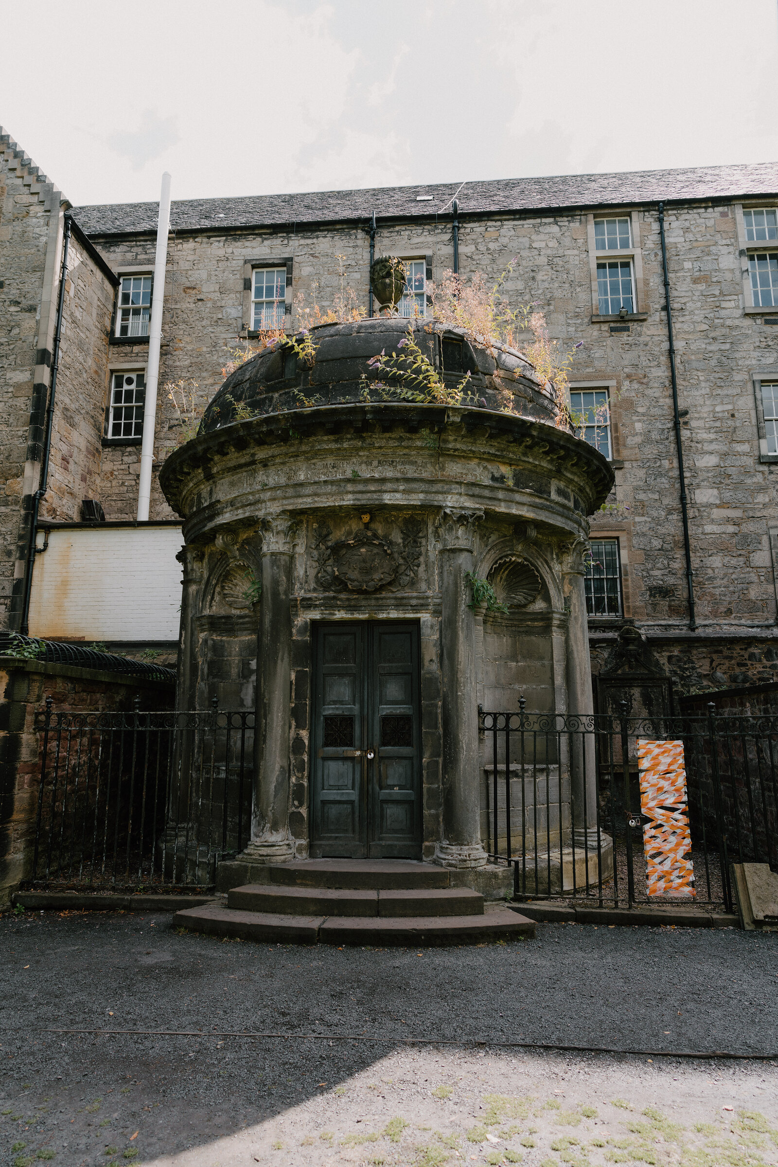 A domed stone mausoleum with iron railings stands in a kirkyard beneath tall tenement buildings.