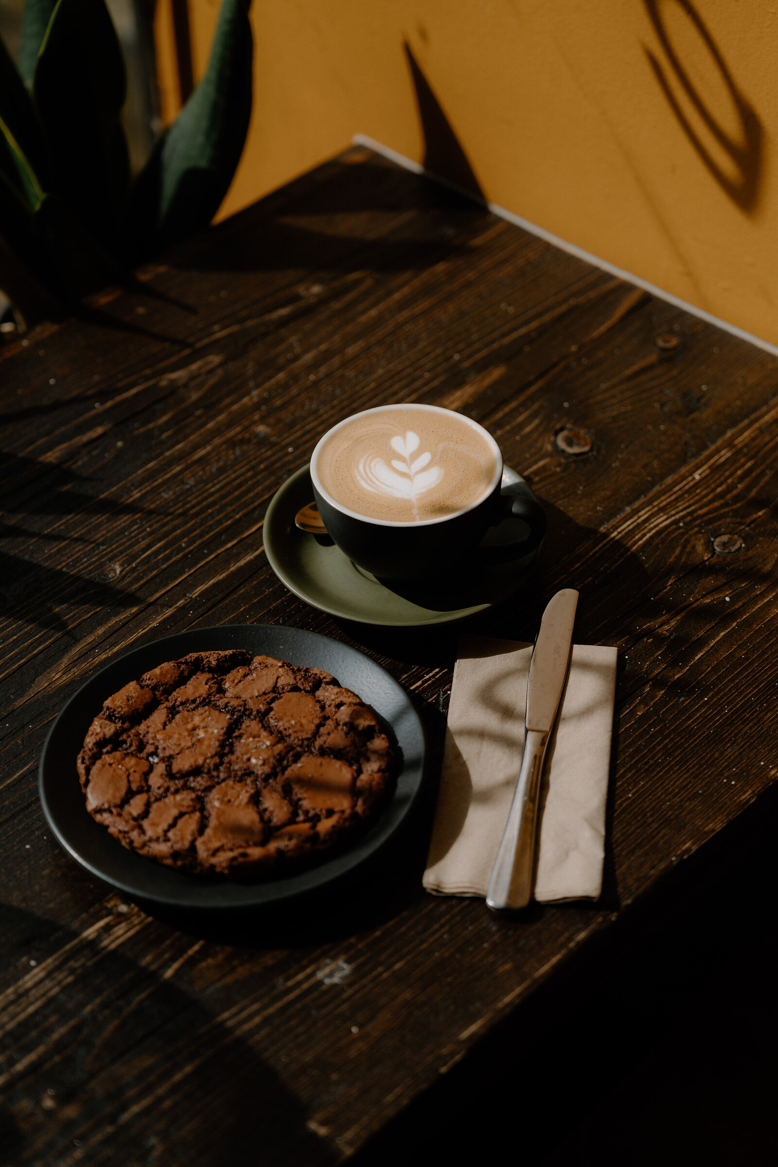 A cappuccino with latte art and a chocolate cookie are served on a sunlit wooden table at grow urban.