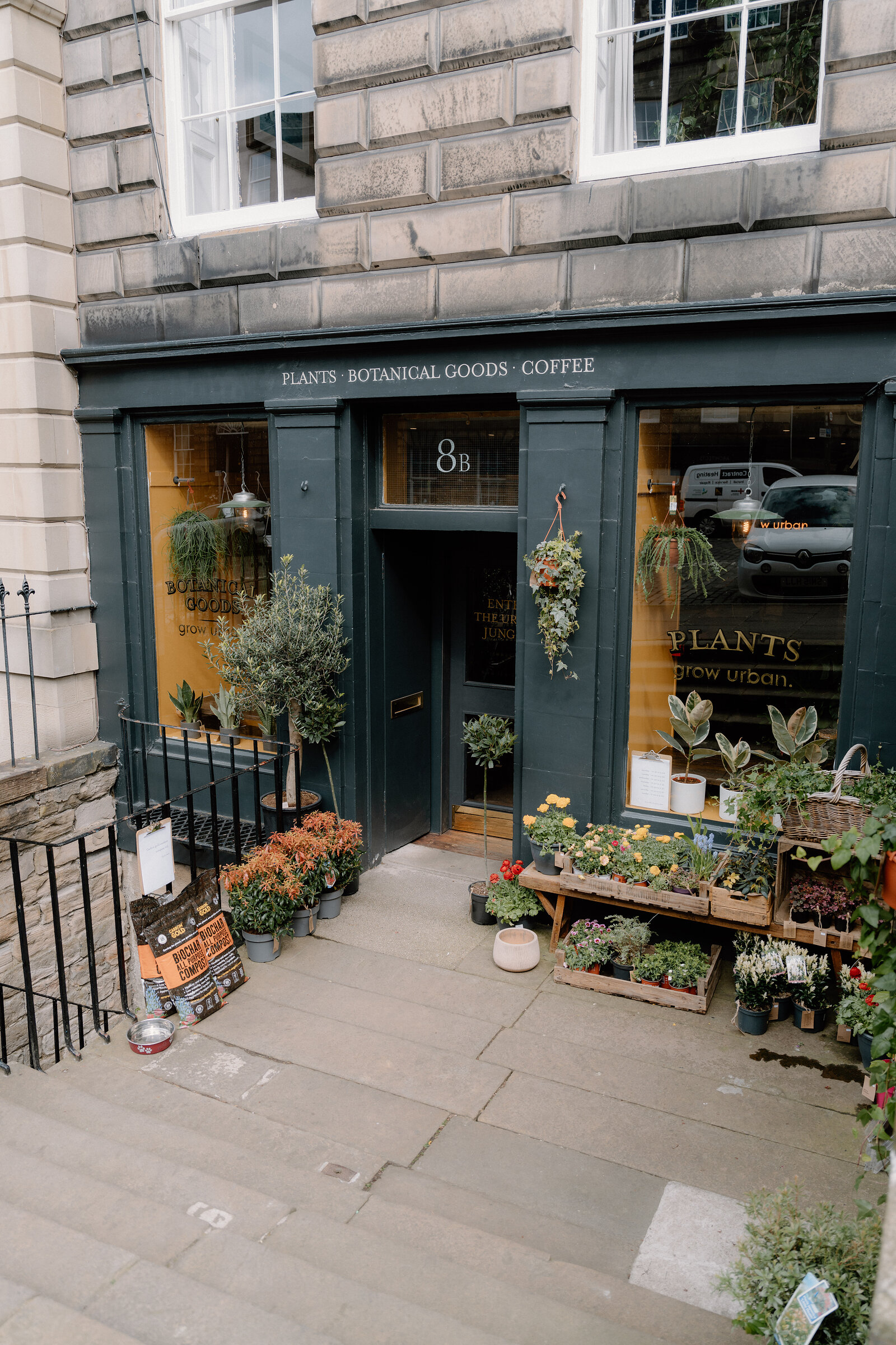 Potted plants and flowers crowd the pavement outside grow urban's shopfront, lettered 'Plants, Botanical Goods, Coffee'.