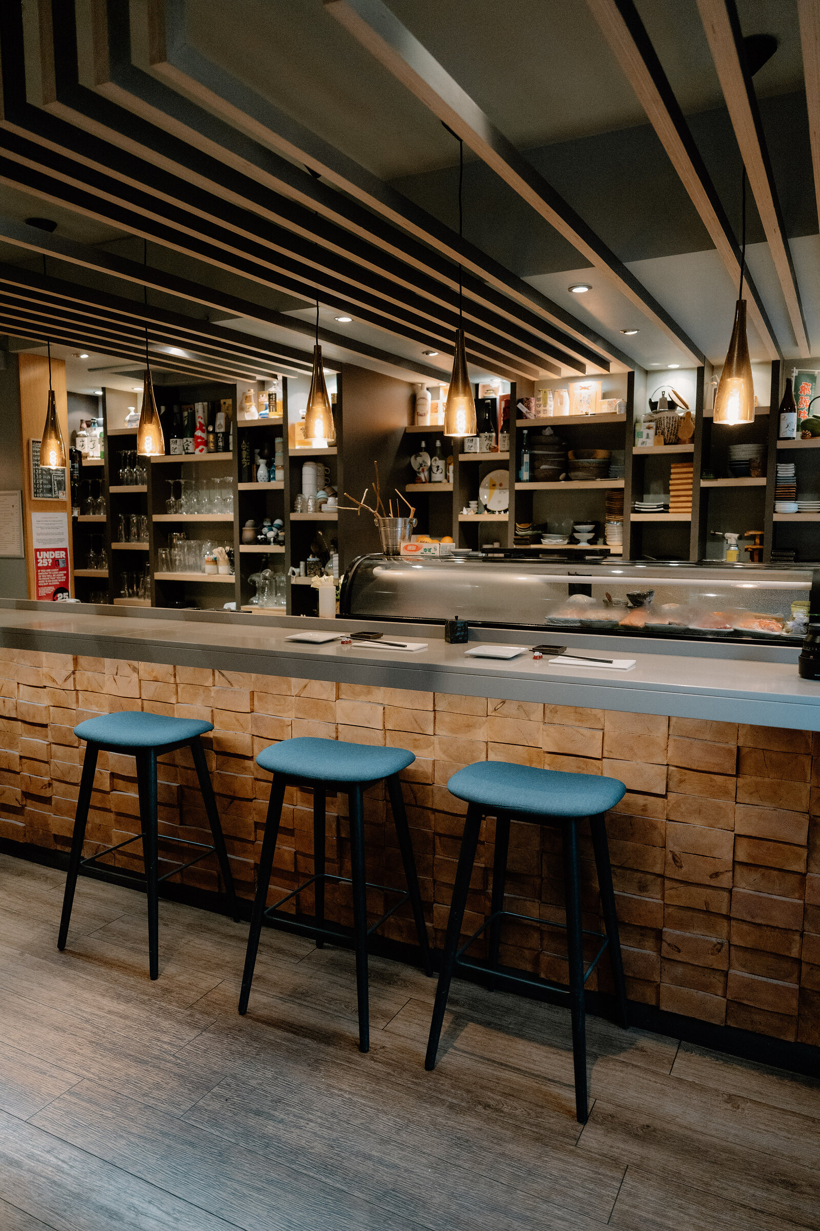 Blue-topped bar stools line a stone-clad sushi counter beneath a slatted wooden ceiling and pendant lights.