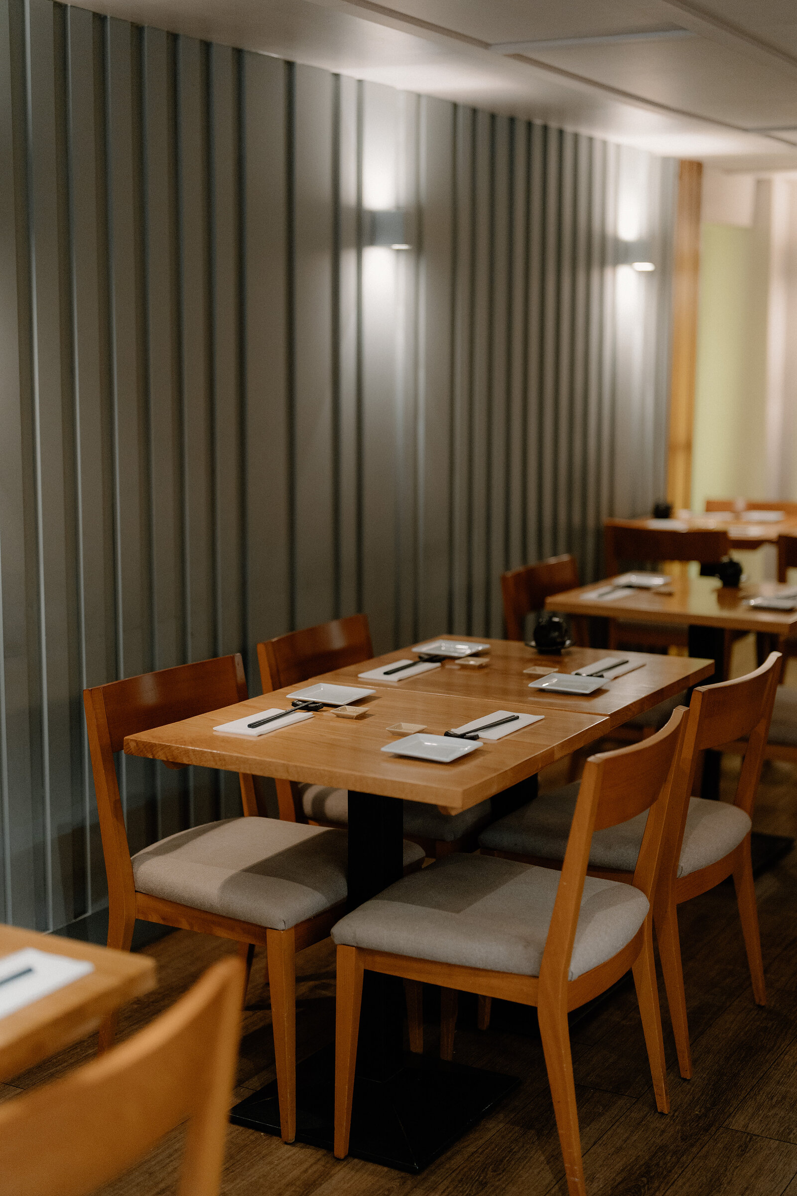 Wooden tables are laid with chopsticks and small plates against a corrugated grey wall in a Japanese restaurant's dining room.