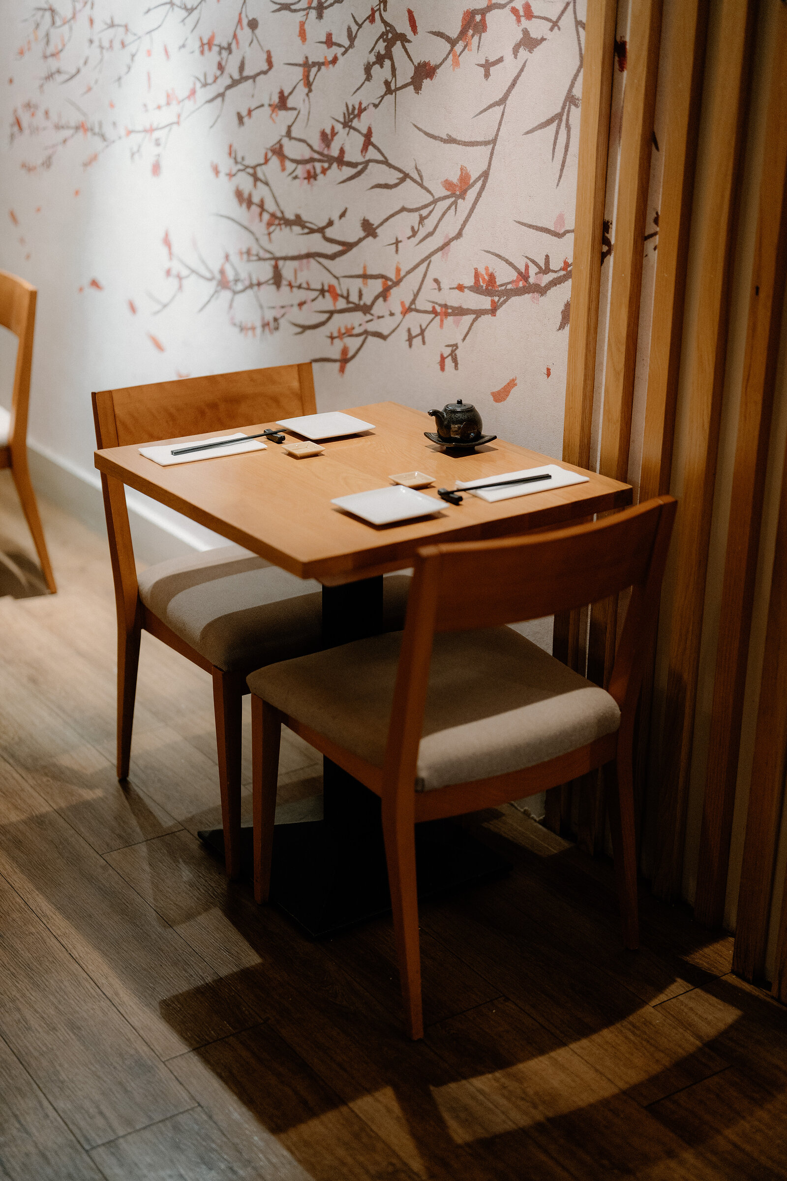 A table set with chopsticks and a small teapot sits beneath a cherry-blossom branch mural in a Japanese restaurant.