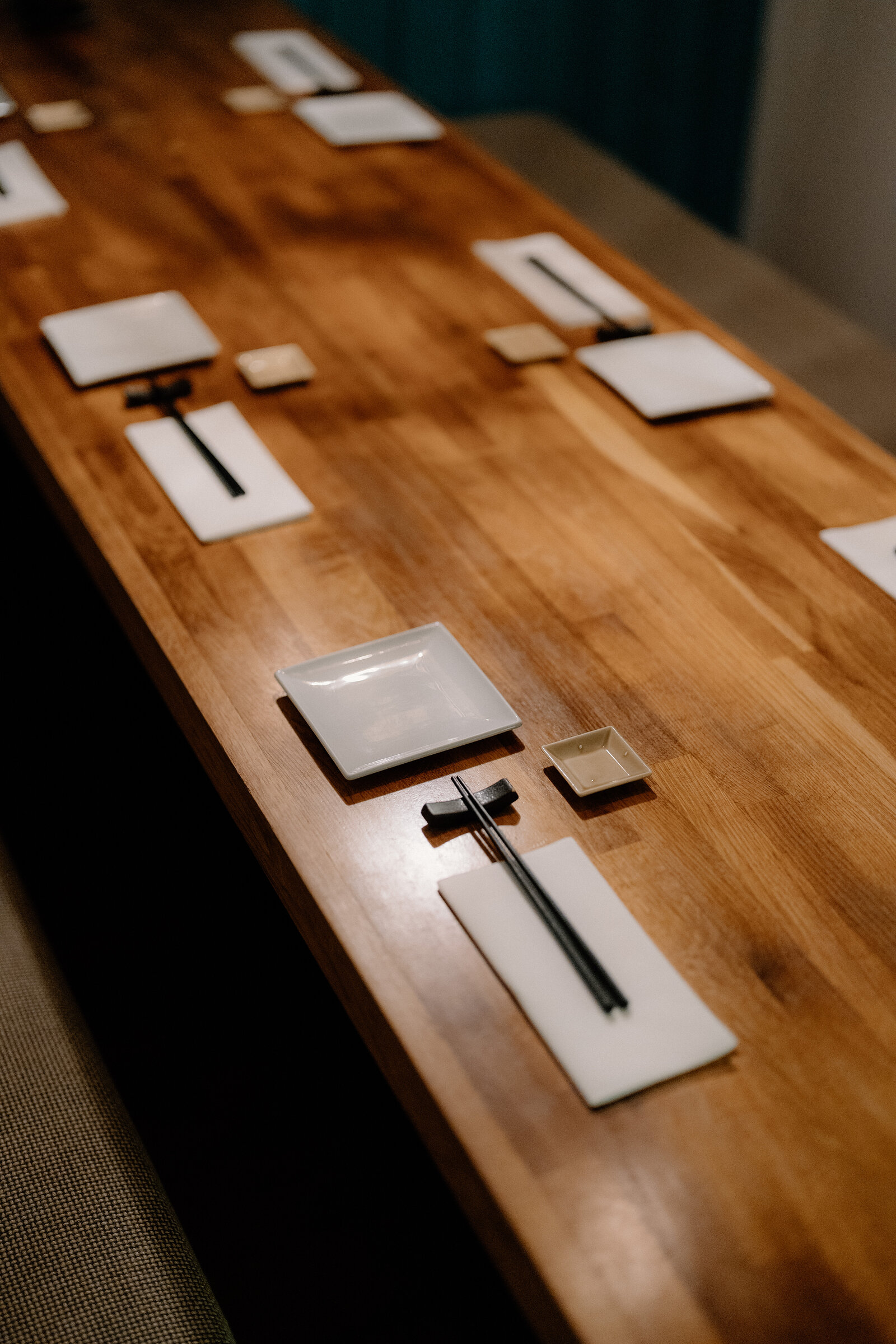 Chopsticks and small dishes are arranged along a long wooden counter in a Japanese restaurant.