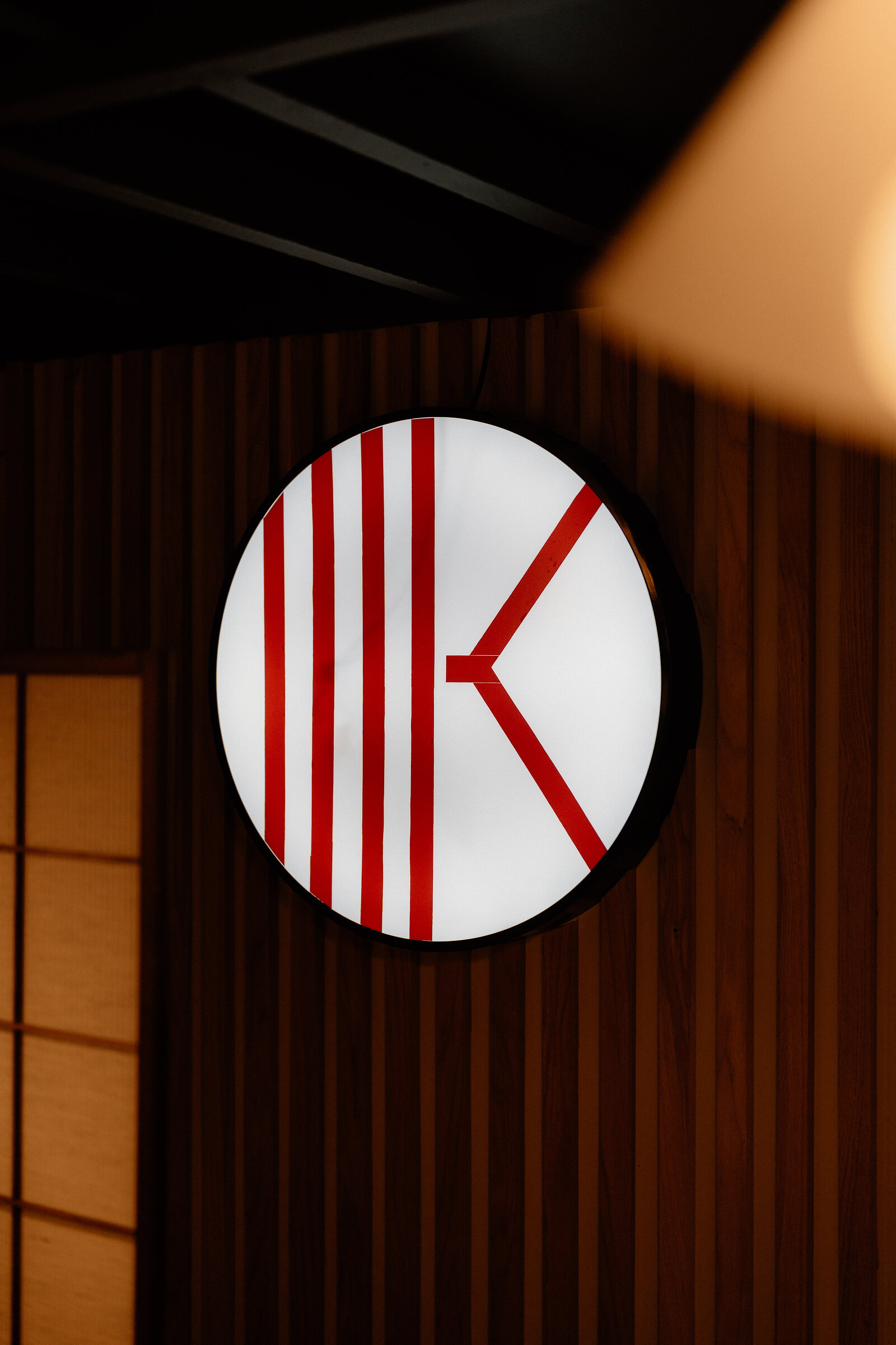 A backlit circular sign with a red letter 'K' logo hangs on a wood-slatted wall beside a paper screen door.