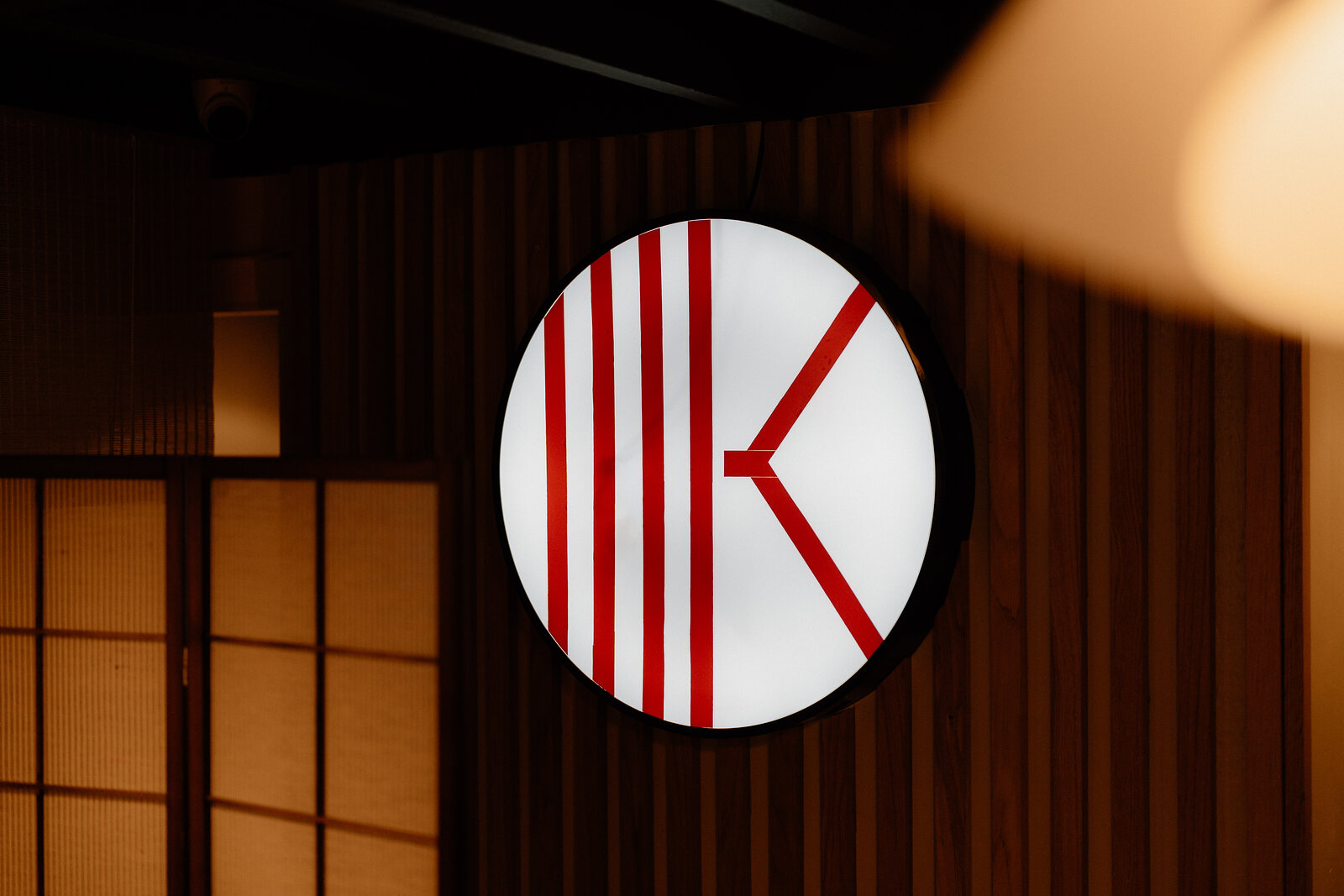 A backlit circular sign with a red letter 'K' logo is mounted on a wood-slatted wall.