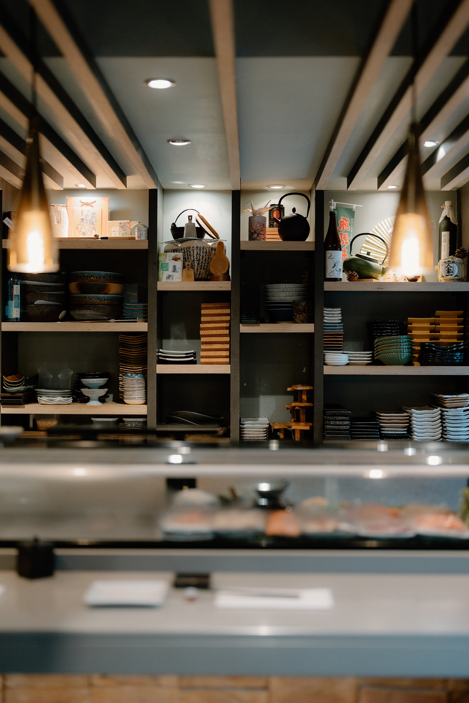 Teapots, bowls and stacked plates fill dark wooden shelving above a sushi counter.