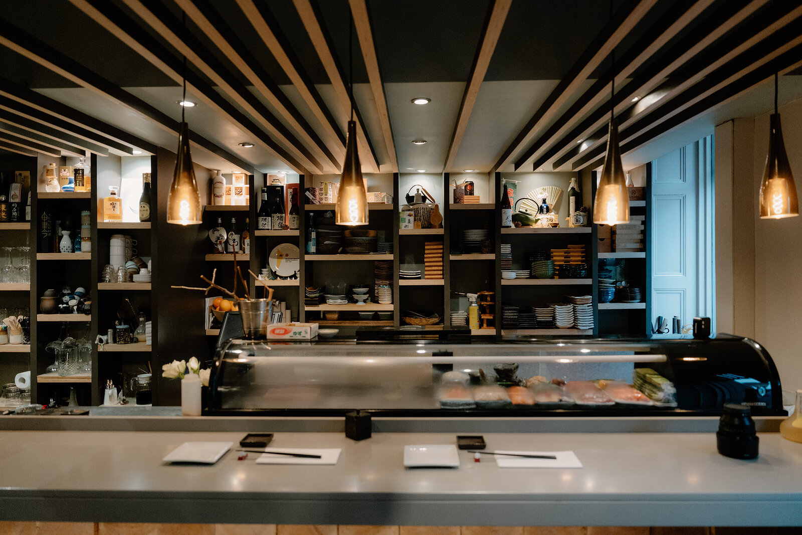 A sushi counter runs beneath a slatted wooden ceiling, lined with plates and glassware on dark shelving.