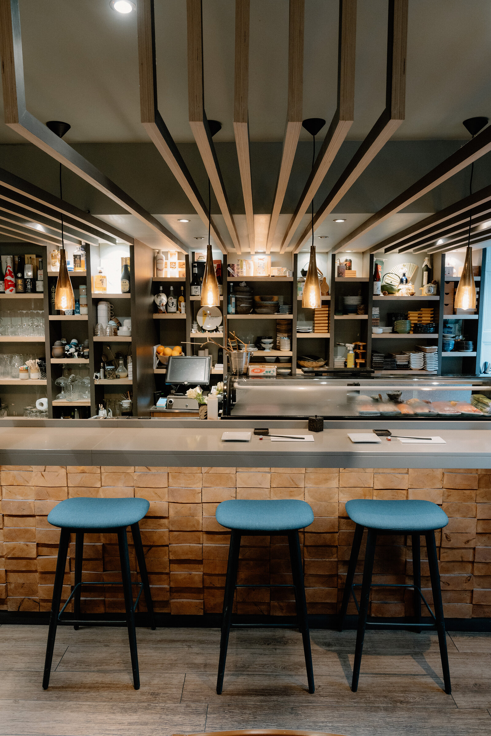 Blue-topped bar stools sit at a brick-clad sushi counter beneath rows of pendant lights.
