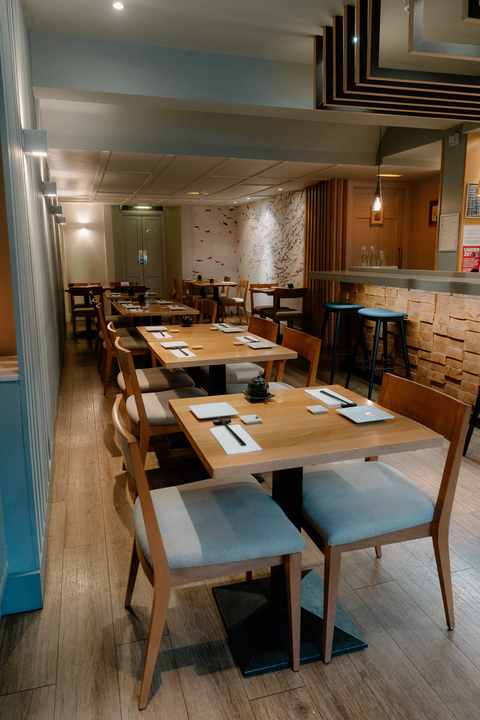 Tables set with chopsticks fill a Japanese restaurant's dining room, with a sushi counter and blue stools visible beyond.