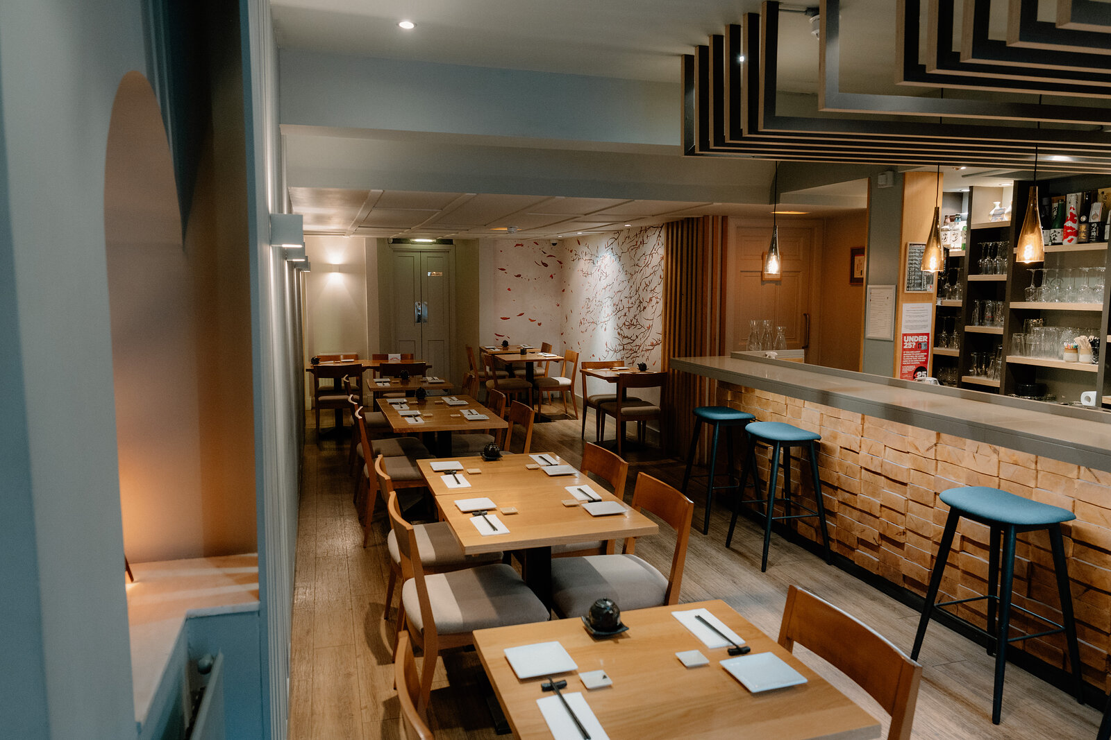 A dining room with wooden tables leads toward a brick-clad sushi counter and blue stools in a Japanese restaurant.