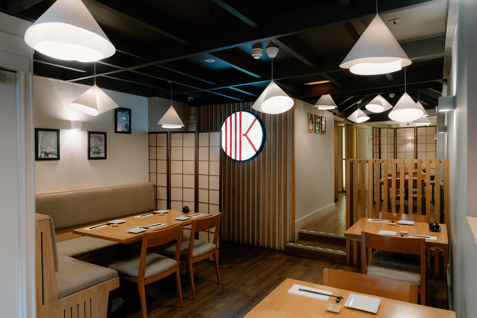 A backlit circular sign with a red letter 'K' logo hangs above dining tables and a paper screen partition.