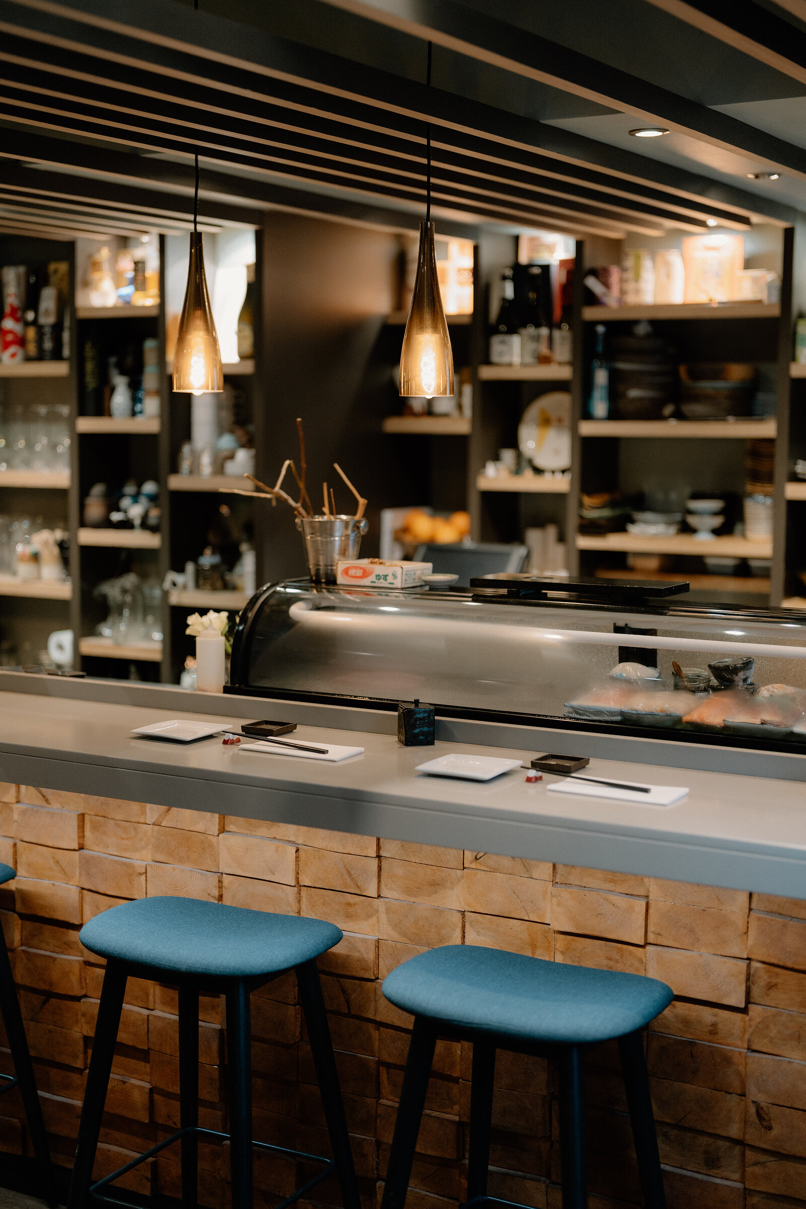 A sushi counter holds trays of fish beneath pendant lights, with blue stools set along its brick-clad front.