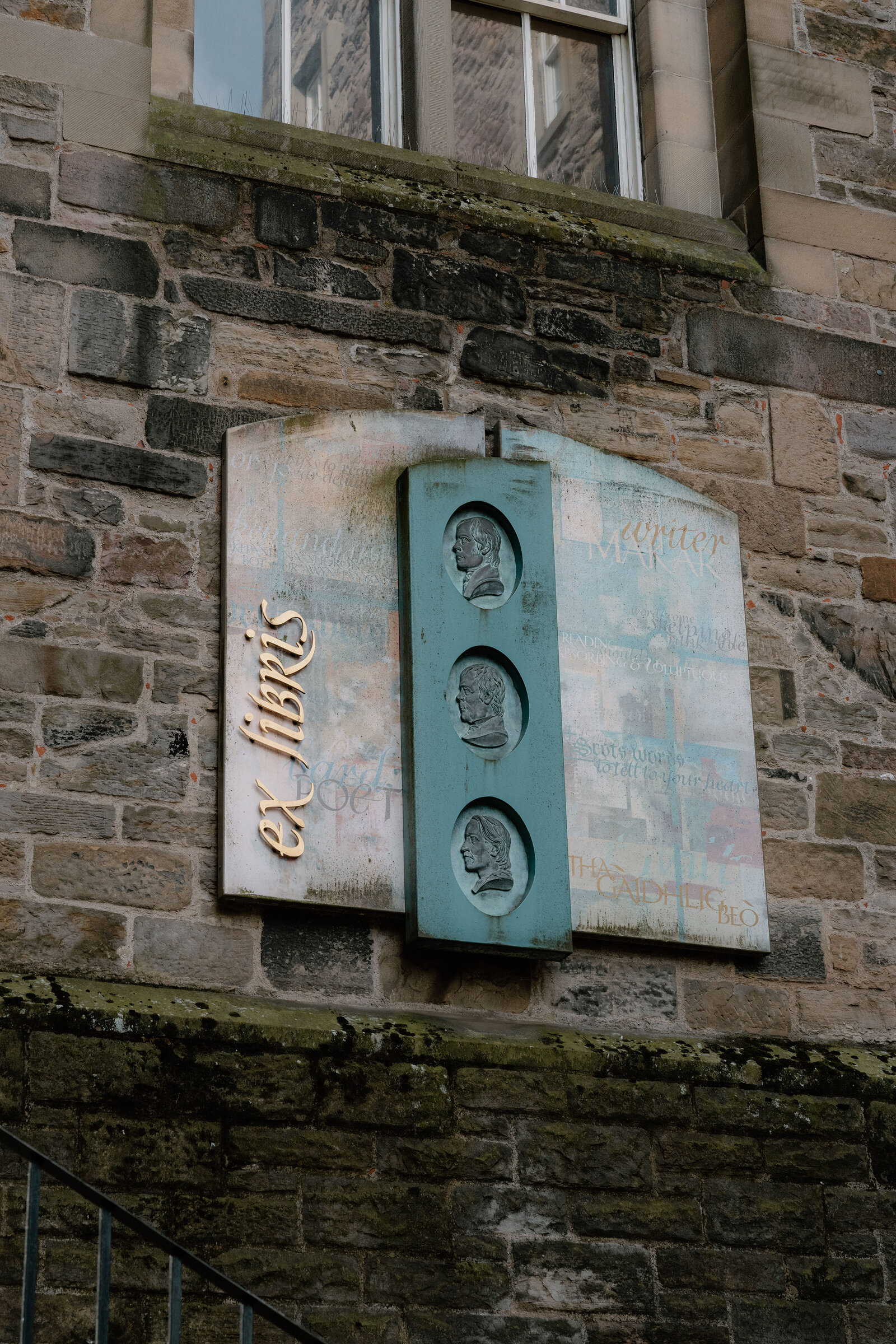 A weathered plaque reading 'ex libris' with three carved portrait medallions is mounted on a stone wall.