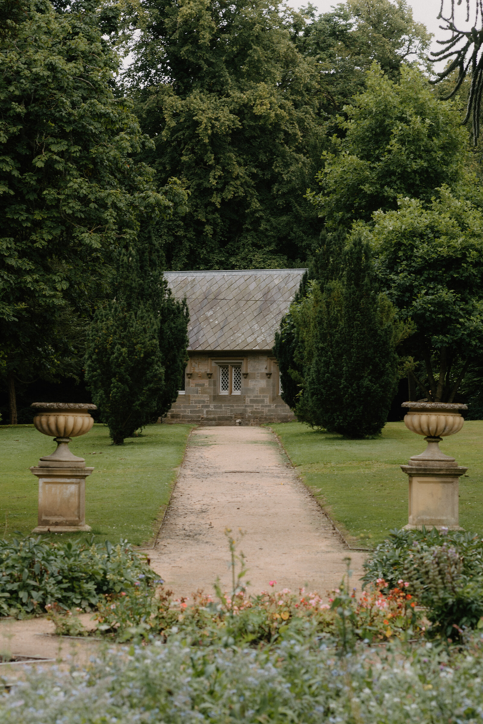 A stone garden pavilion stands at the end of a gravel path flanked by clipped conifers and stone urns.