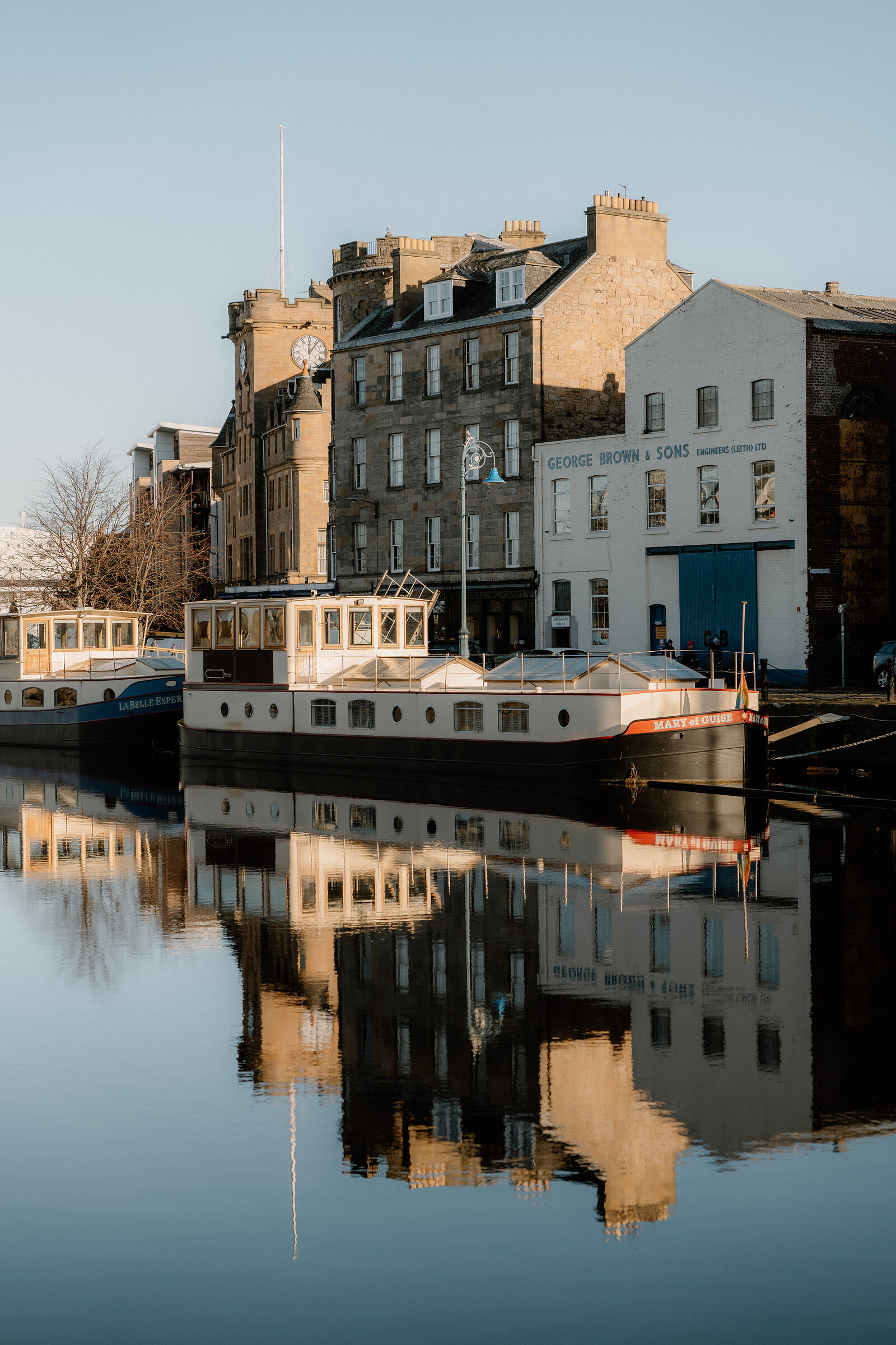 Houseboats are moored below a clock tower and the 'George Brown & Sons' building on Leith's waterfront, reflected in still water.