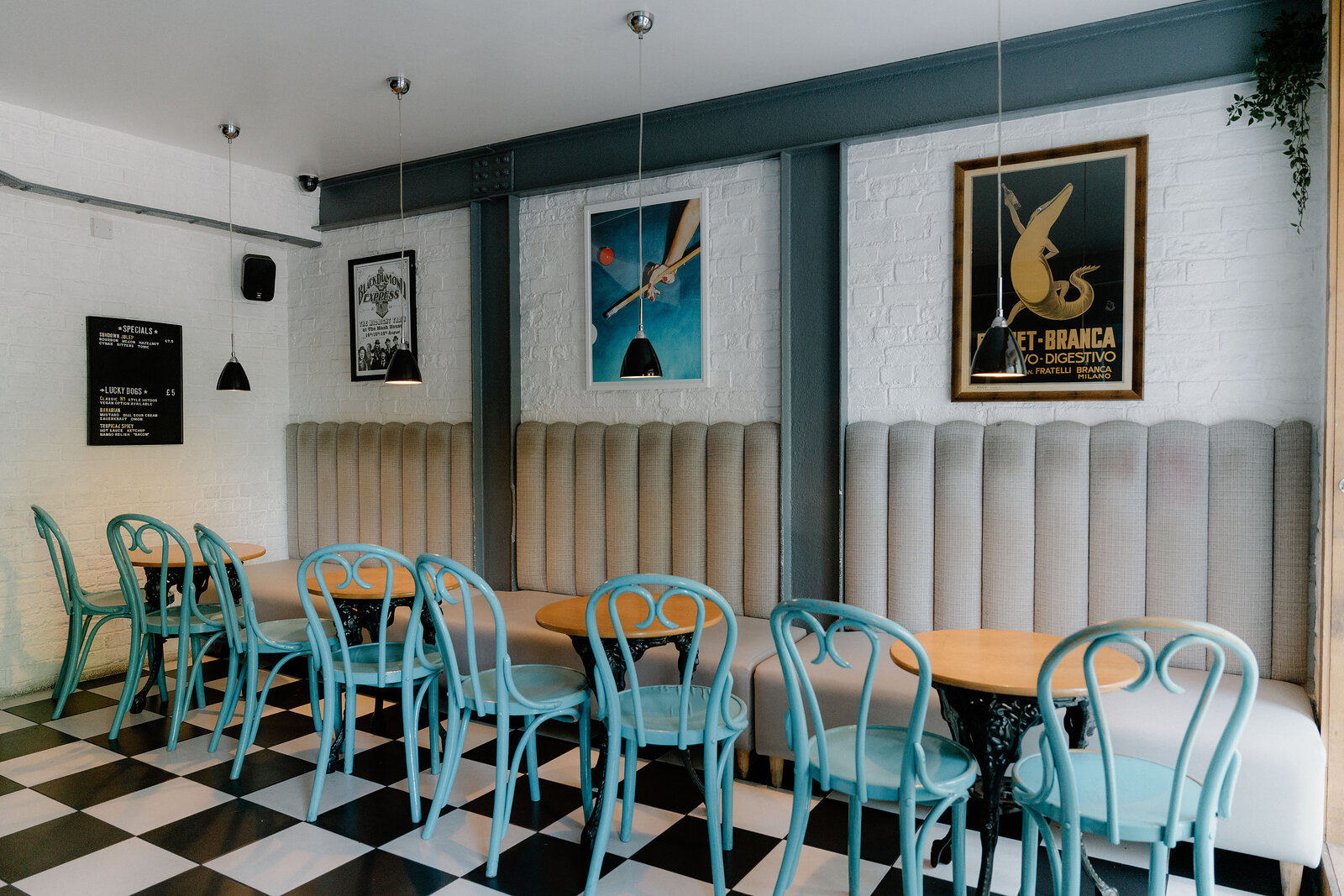 Teal bentwood chairs and beige banquette seating sit beneath vintage aperitif posters on a checkerboard floor in an Edinburgh bar.