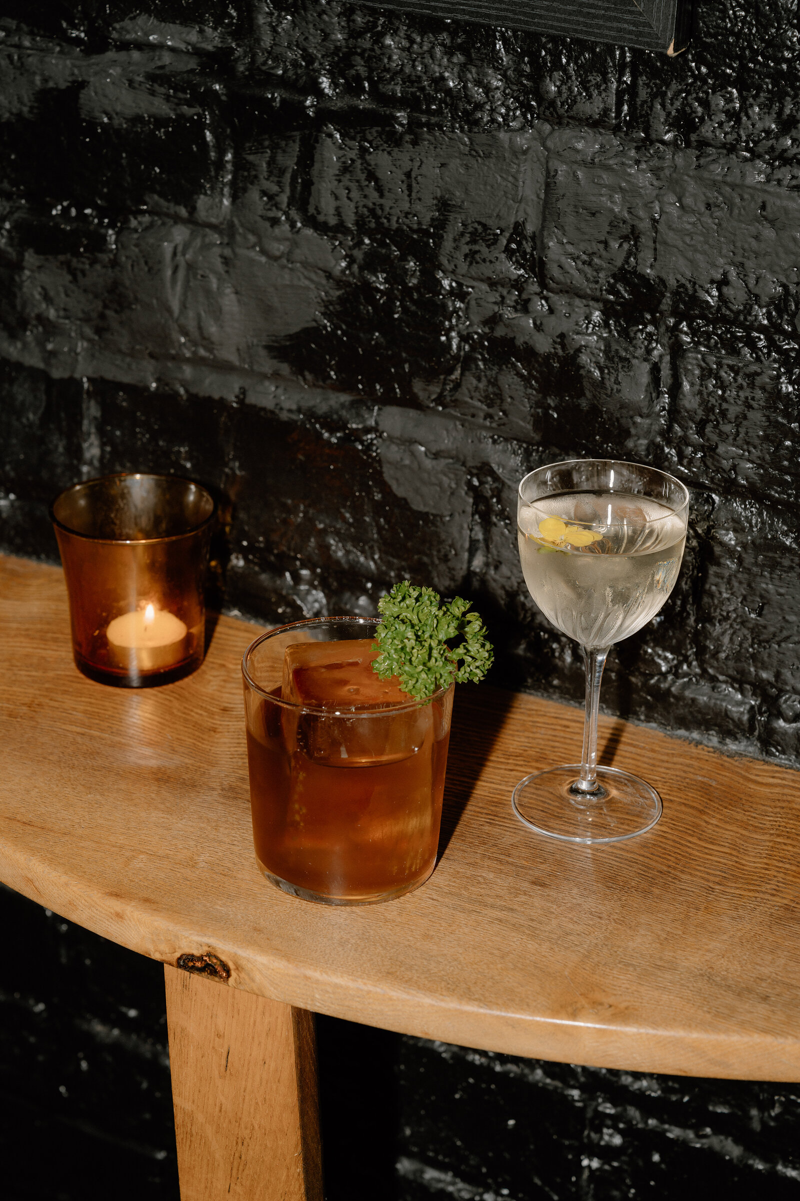 A candle, an Old Fashioned and a martini are arranged on a table against a black brick wall in an Edinburgh bar.