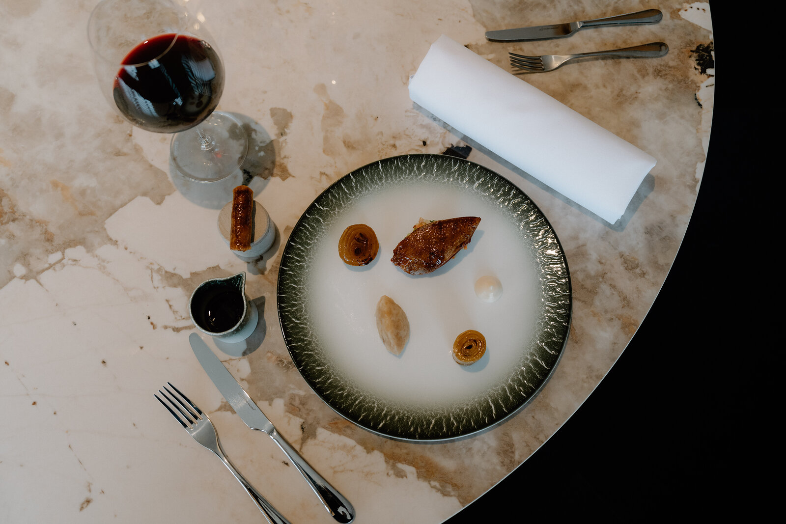 A seared meat course is plated beside a glass of red wine and a rolled napkin on a marble table in an Edinburgh restaurant.