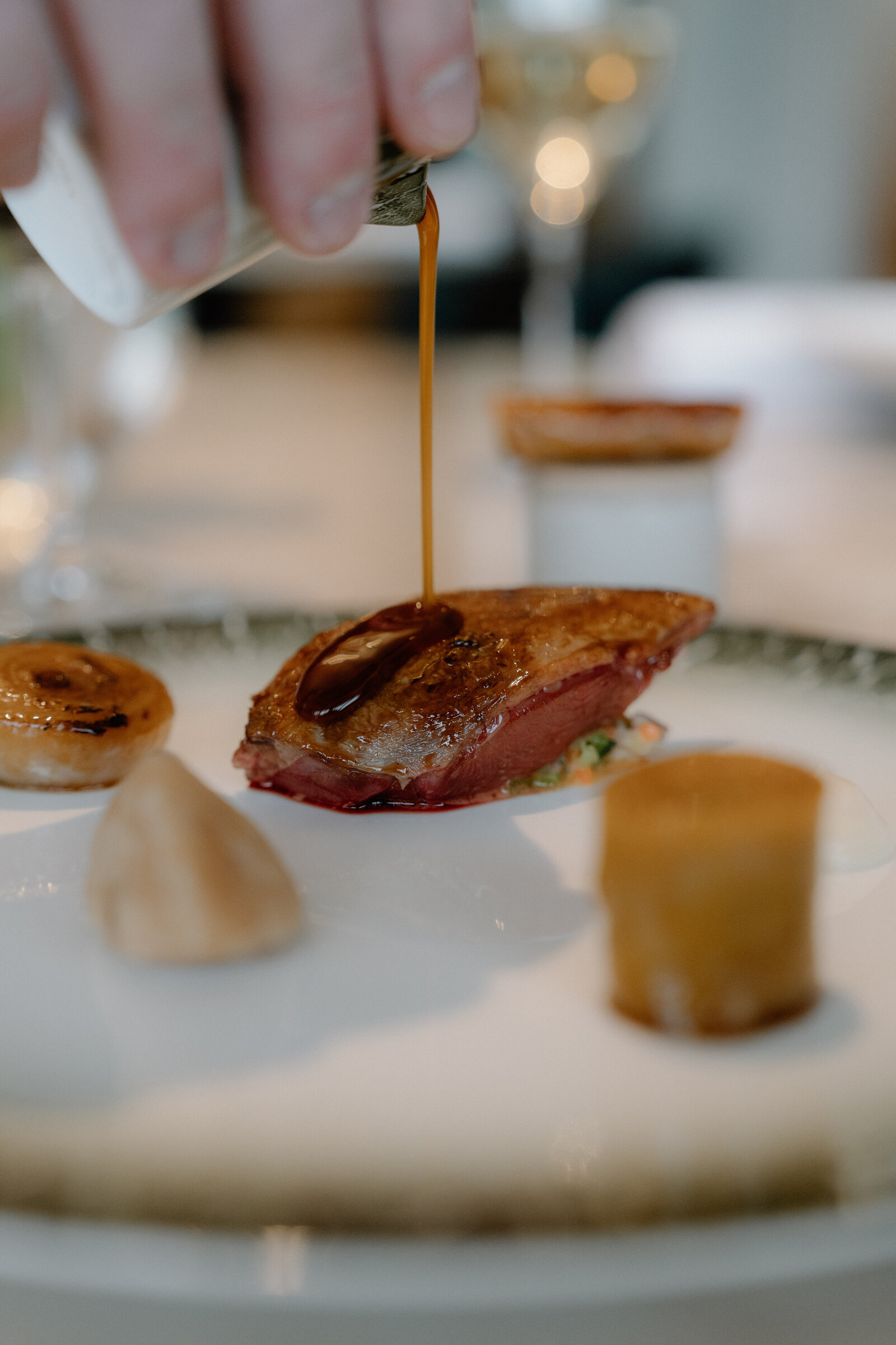 A hand pours sauce over a plated meat course beside glasses of wine in an Edinburgh restaurant.
