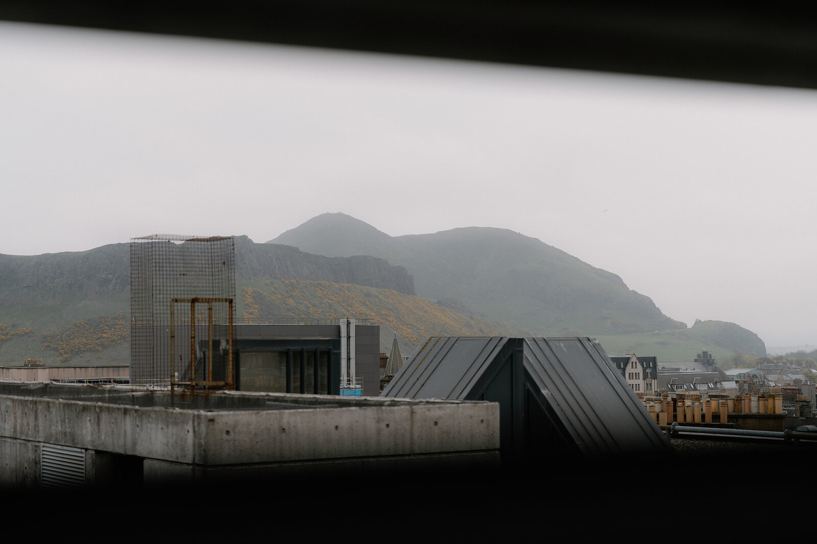 Salisbury Crags rise beyond a modern concrete rooftop structure and angular metal roof vents in Edinburgh.