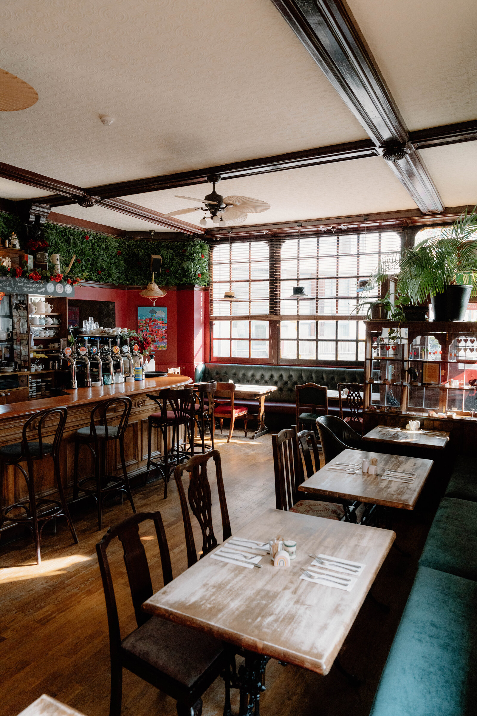 A green banquette and dining tables sit beneath a foliage-trimmed ceiling in a pub, a bar visible beyond.