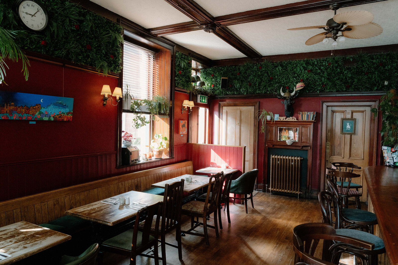 Foliage-trimmed red walls surround a fireplace and dining tables in a pub, a stag's head mounted above the mantel.