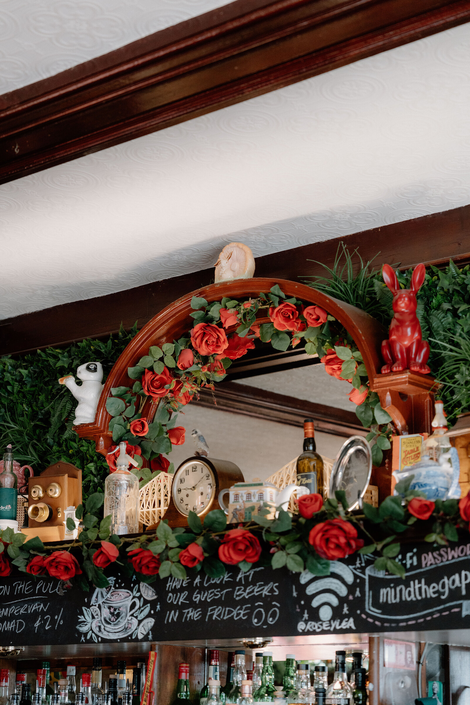 A rose garland frames a mirror above a mantel crowded with ornaments and a red rabbit statue, a chalkboard listing guest beers below.