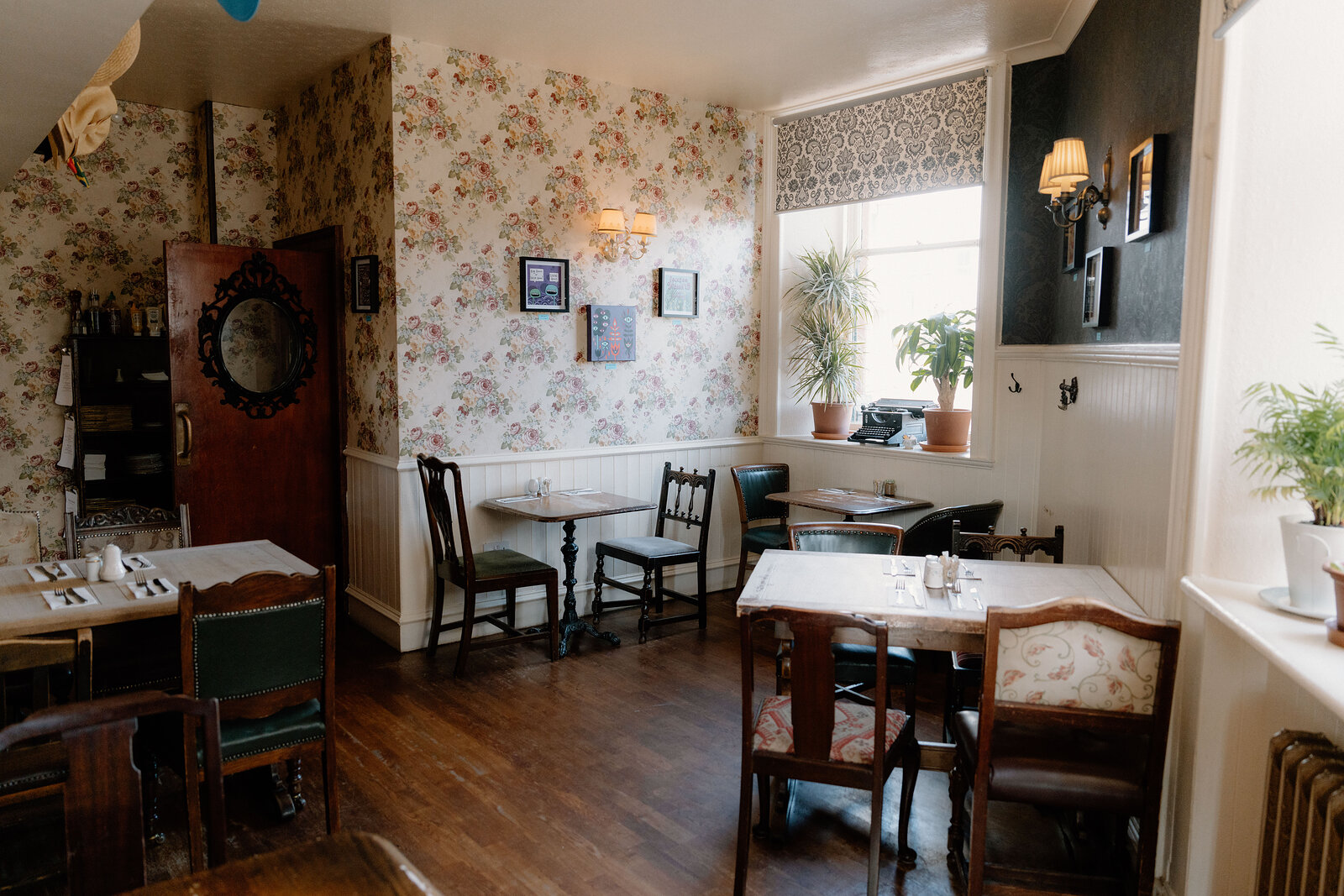 Floral wallpaper and dark wood furniture fill a traditional dining room, a vintage mirror hanging by the door.