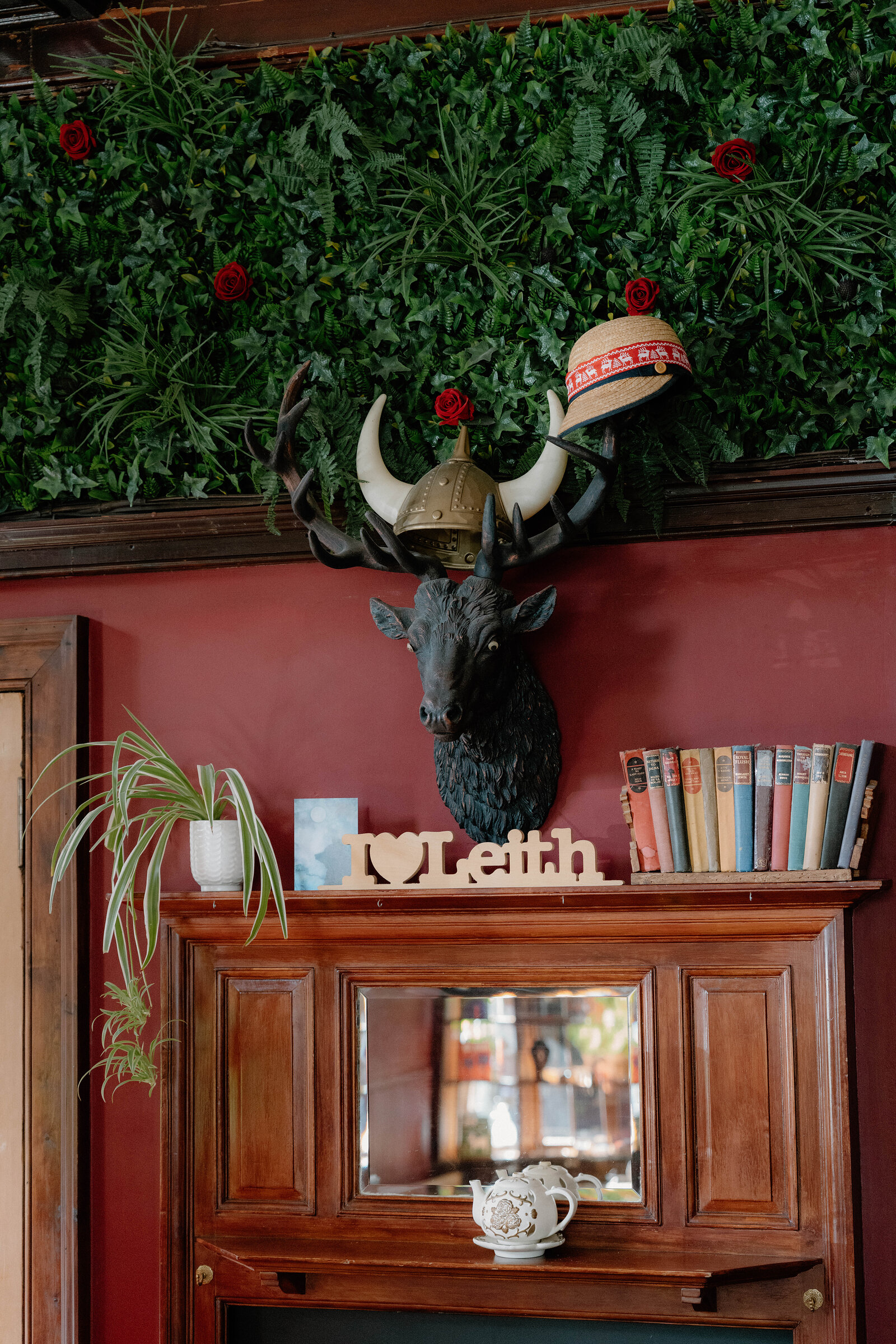 A mounted stag's head wearing a hat sits below a foliage display, a wooden sign reading 'I love Leith' on the mantel beside books.