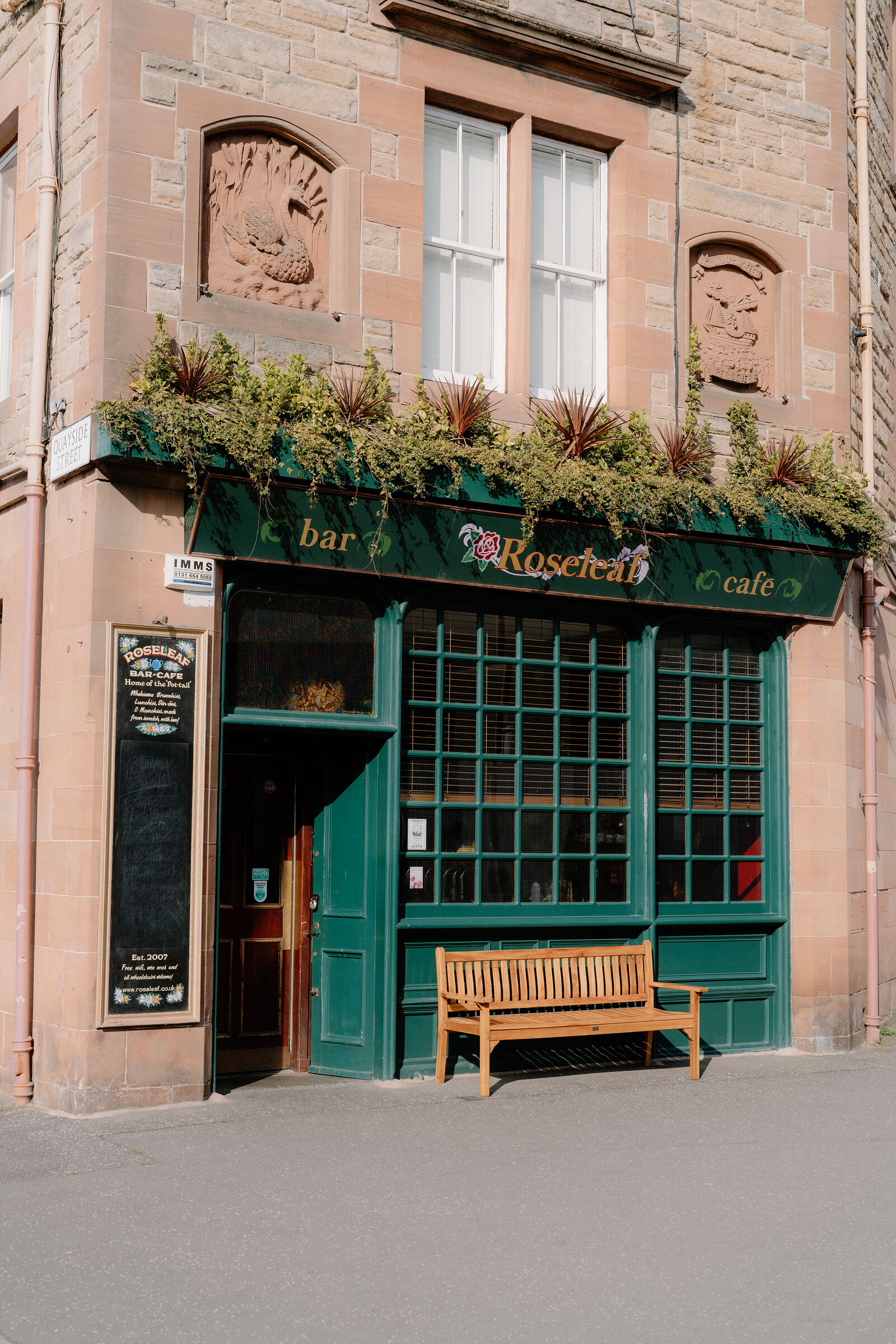 Rosecleft Bar and Cafe's green corner shopfront sits beneath a sandstone tenement, a wooden bench outside.