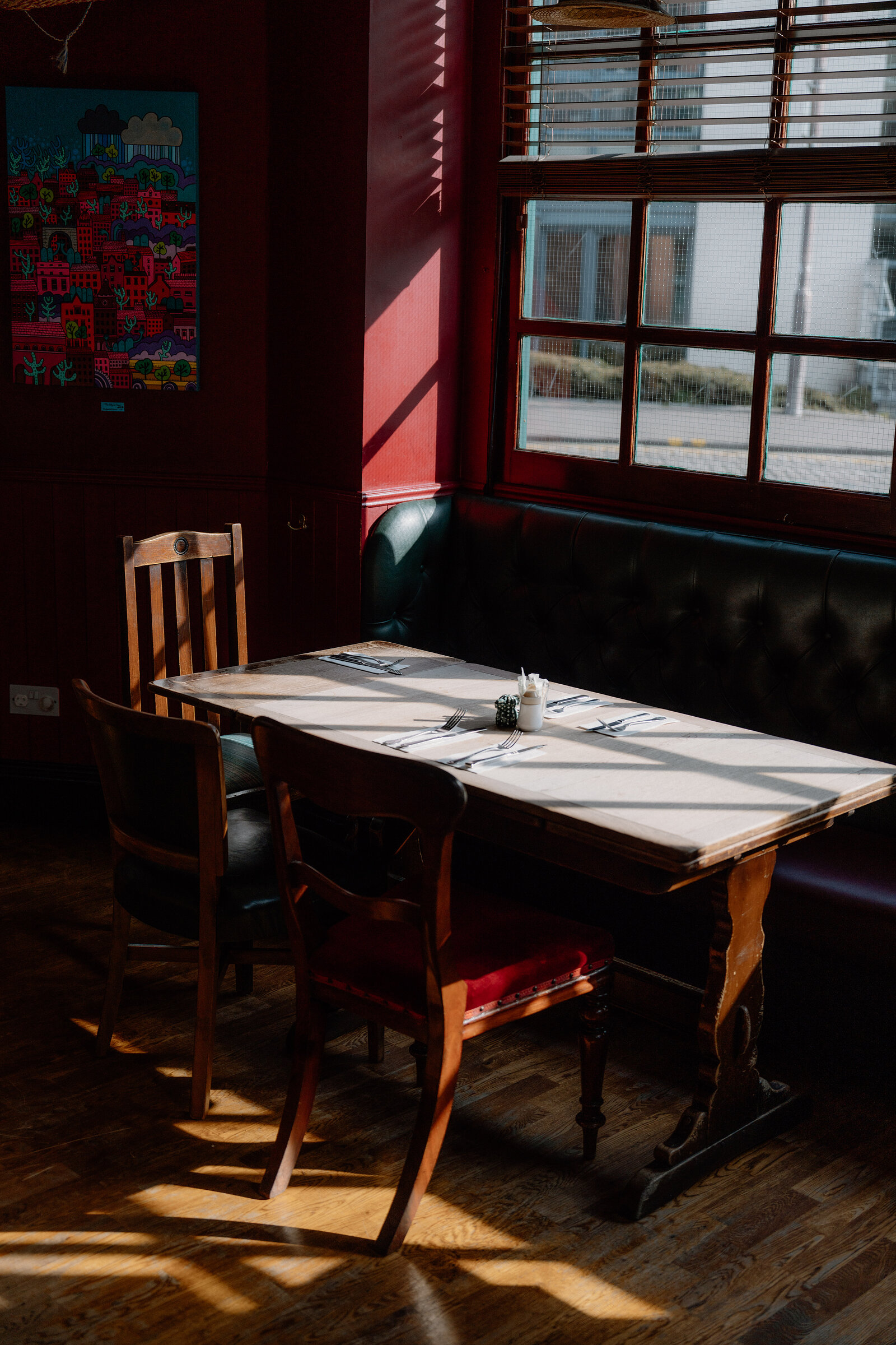 A colourful cityscape print hangs on a red wall beside a wooden table set with cutlery in a restaurant booth.
