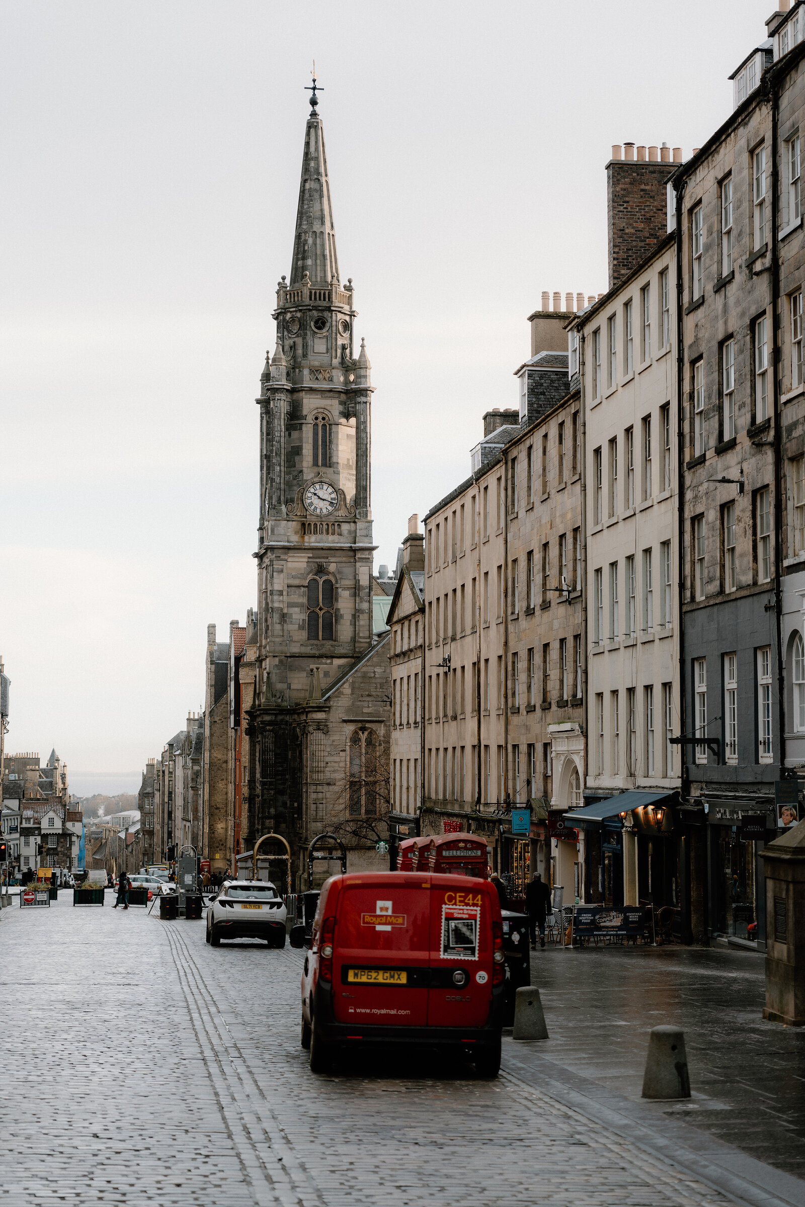 A red postal van is parked on the cobbled Royal Mile beneath the Tron Kirk's clock tower and spire.