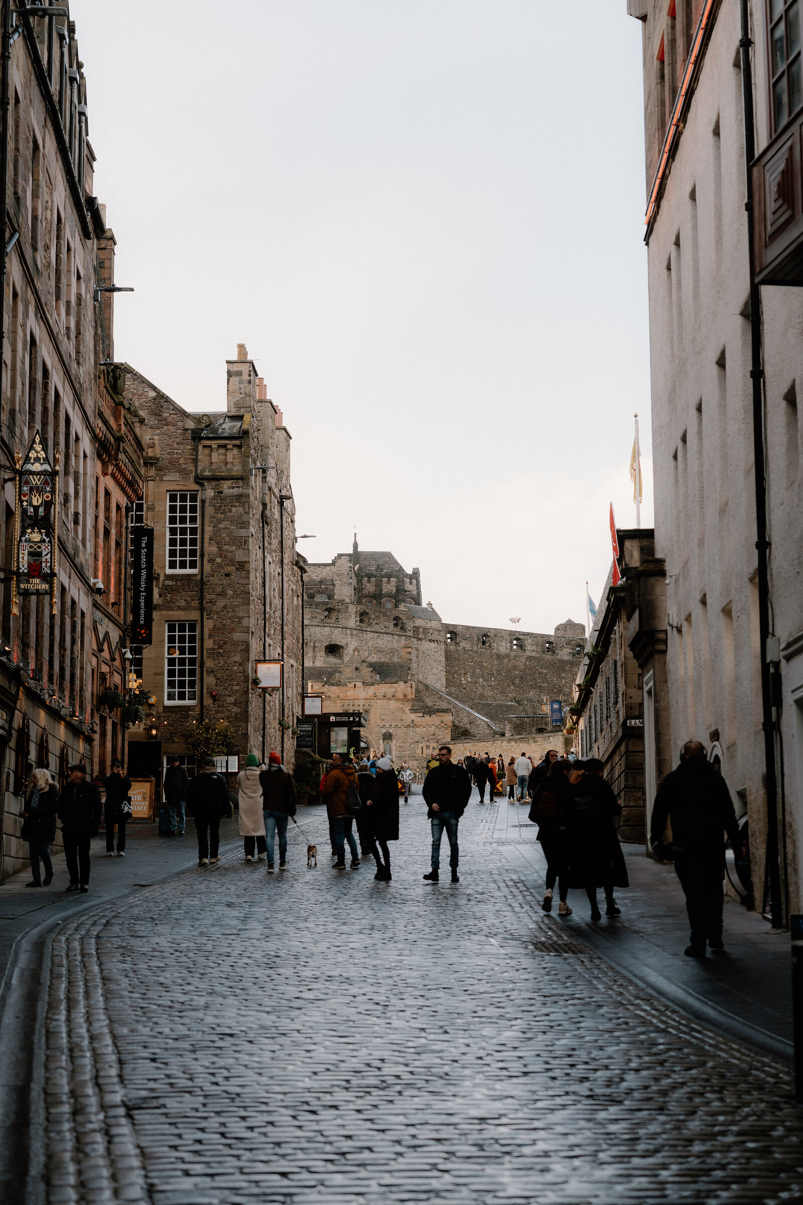 Pedestrians walk a cobbled Royal Mile street toward Edinburgh Castle's ramparts.